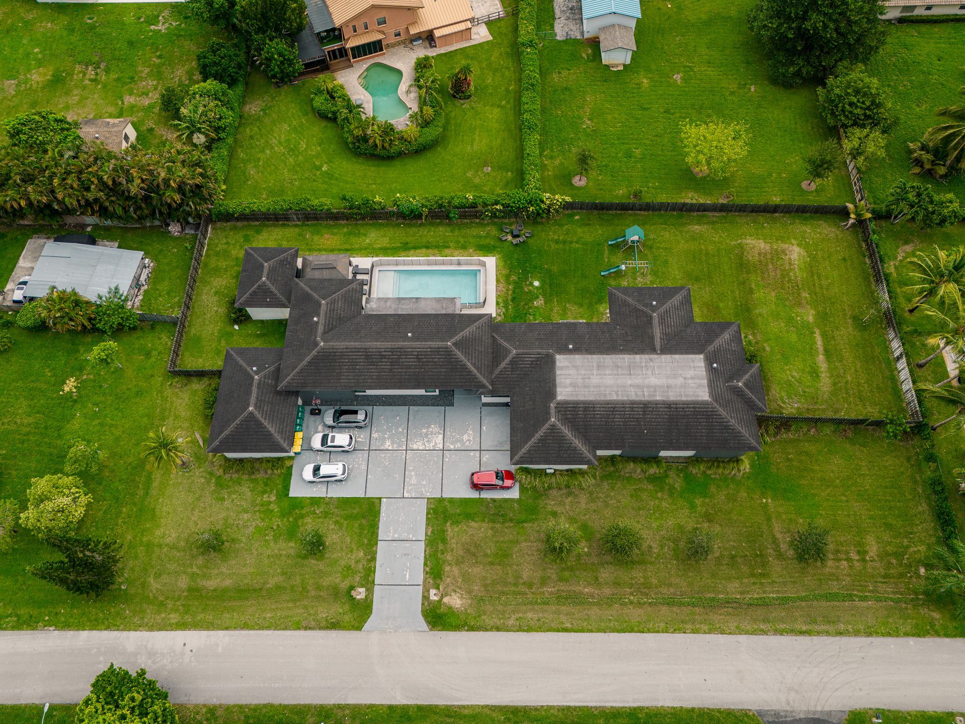 Vista aérea de una casa grande con piscina, vehículos en la entrada y un gran patio cubierto de césped, rodeado por una cerca.