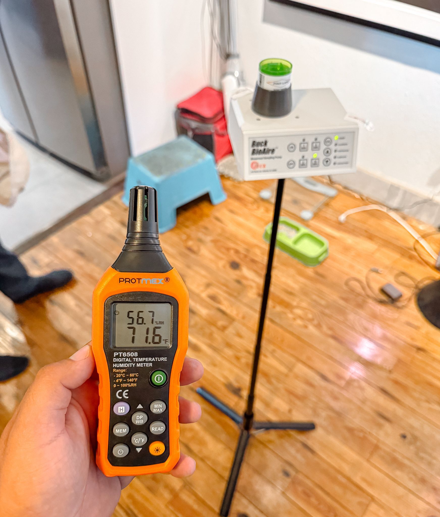 Una persona sostiene un medidor ambiental naranja. Un dispositivo de monitoreo blanco se encuentra en un soporte cercano.