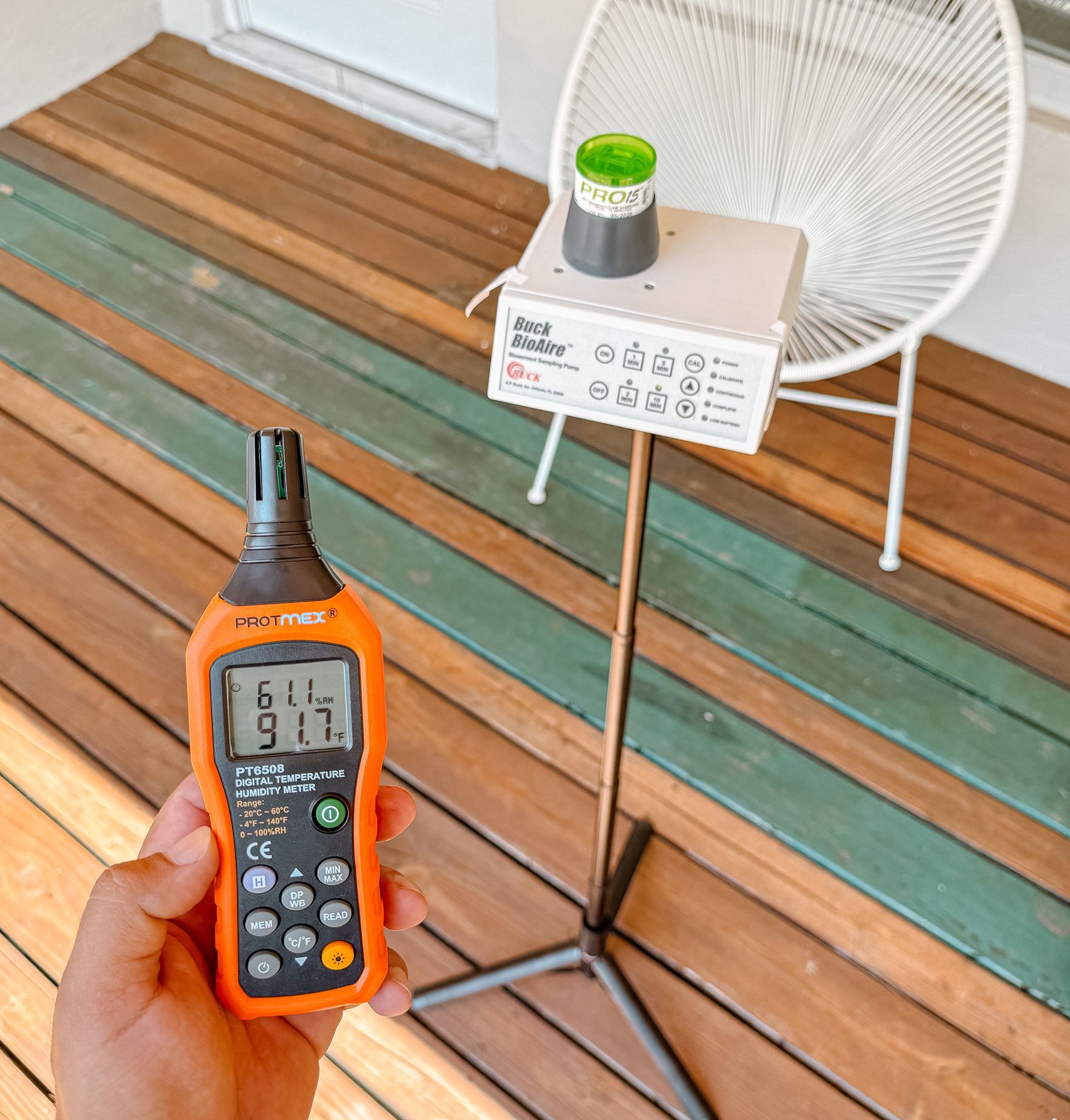 Mano sosteniendo un medidor digital naranja que marca 91.7. Junto a él hay una caja blanca con botones, un soporte y una plataforma de madera.