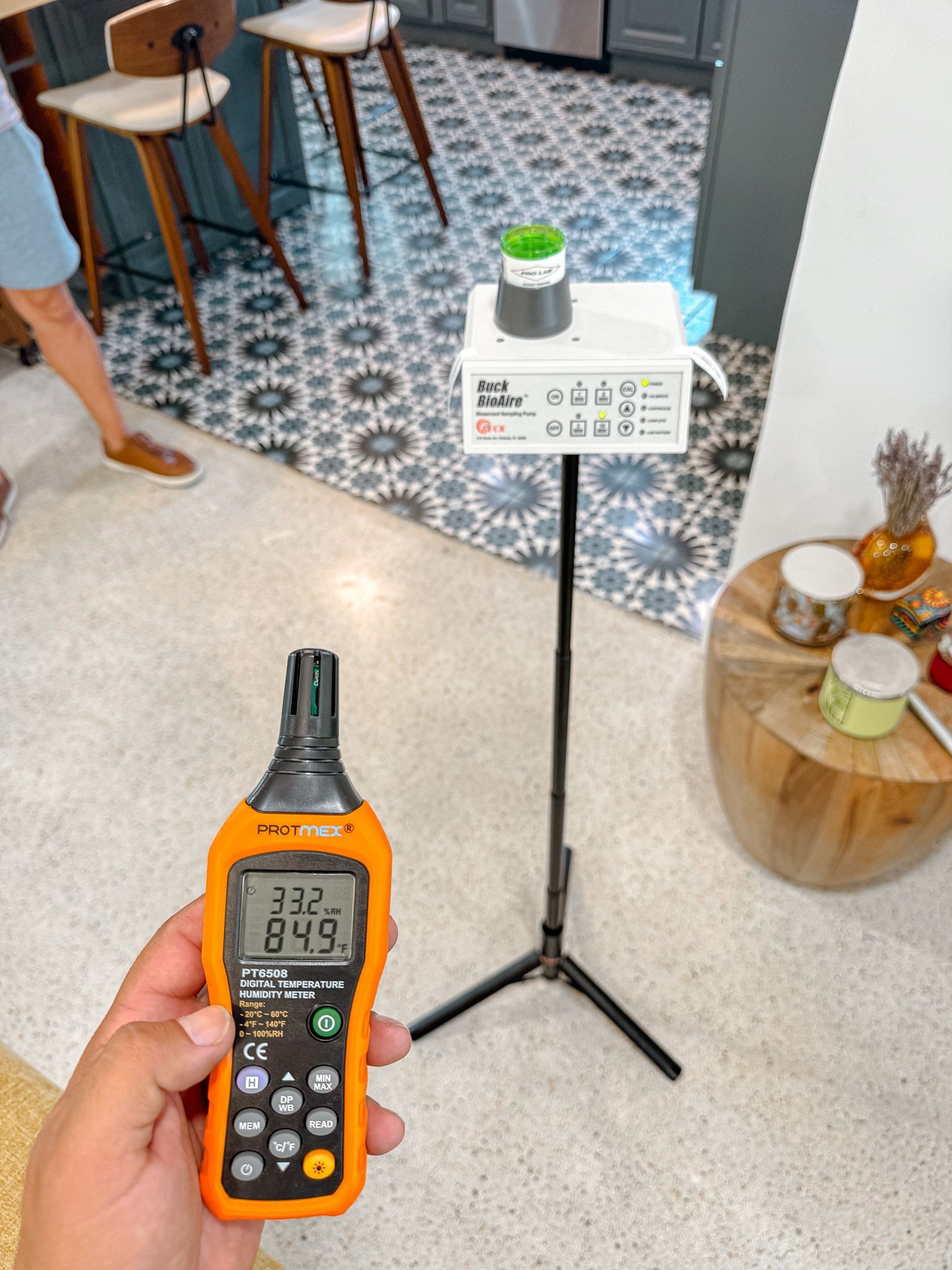 Persona sosteniendo un medidor de humedad digital, con un dispositivo de prueba en un trípode en una habitación.