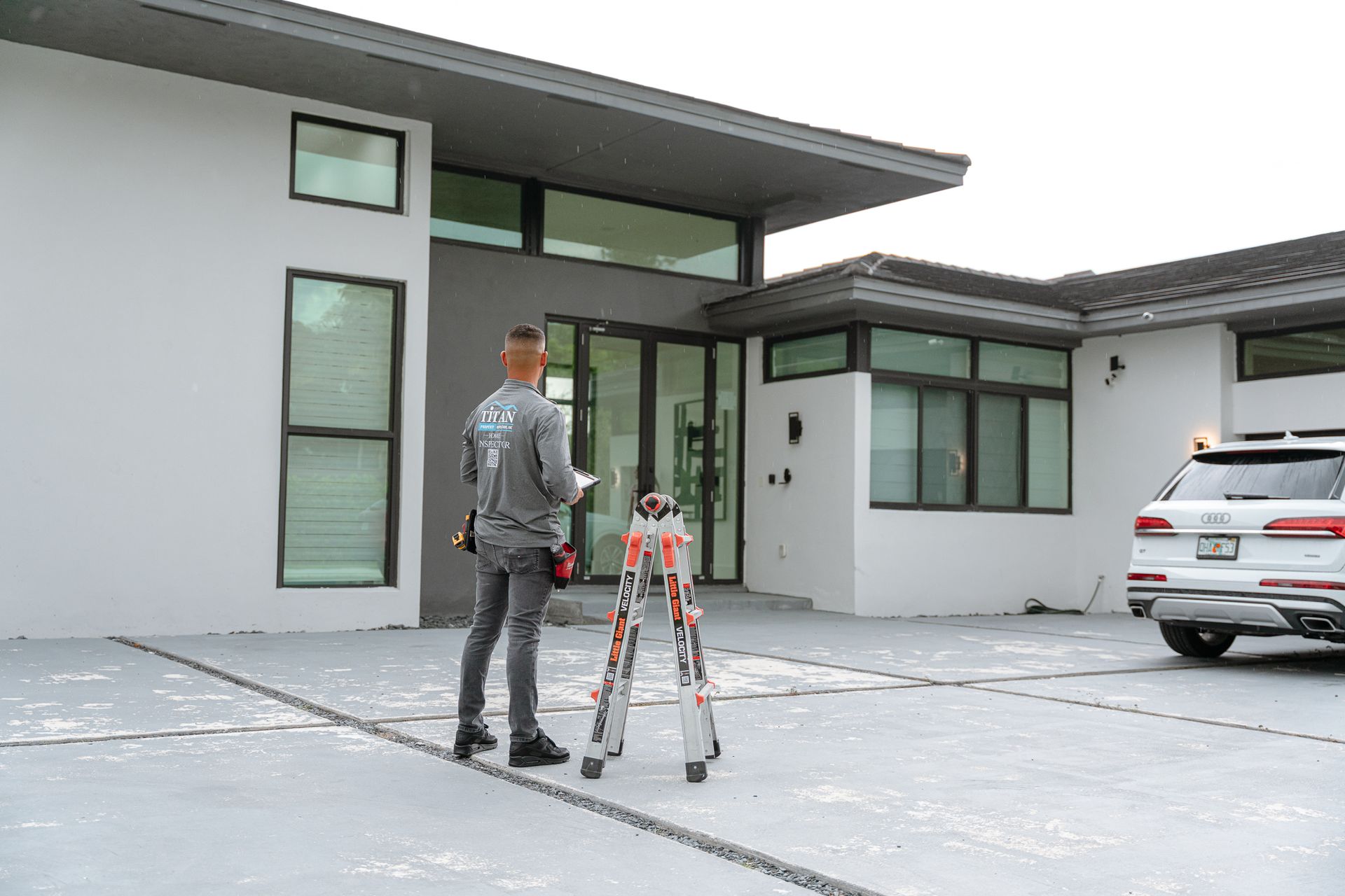Una persona parada frente a una casa blanca moderna, sosteniendo un portapapeles, con un auto estacionado cerca.