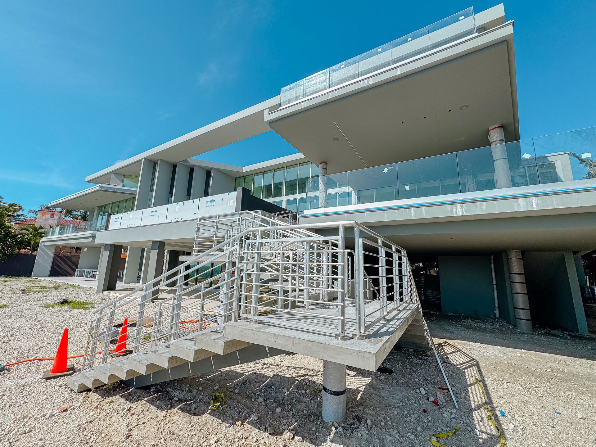 Casa moderna en construcción con balcones de cristal, soportes de hormigón y escalera. Cielo azul brillante.