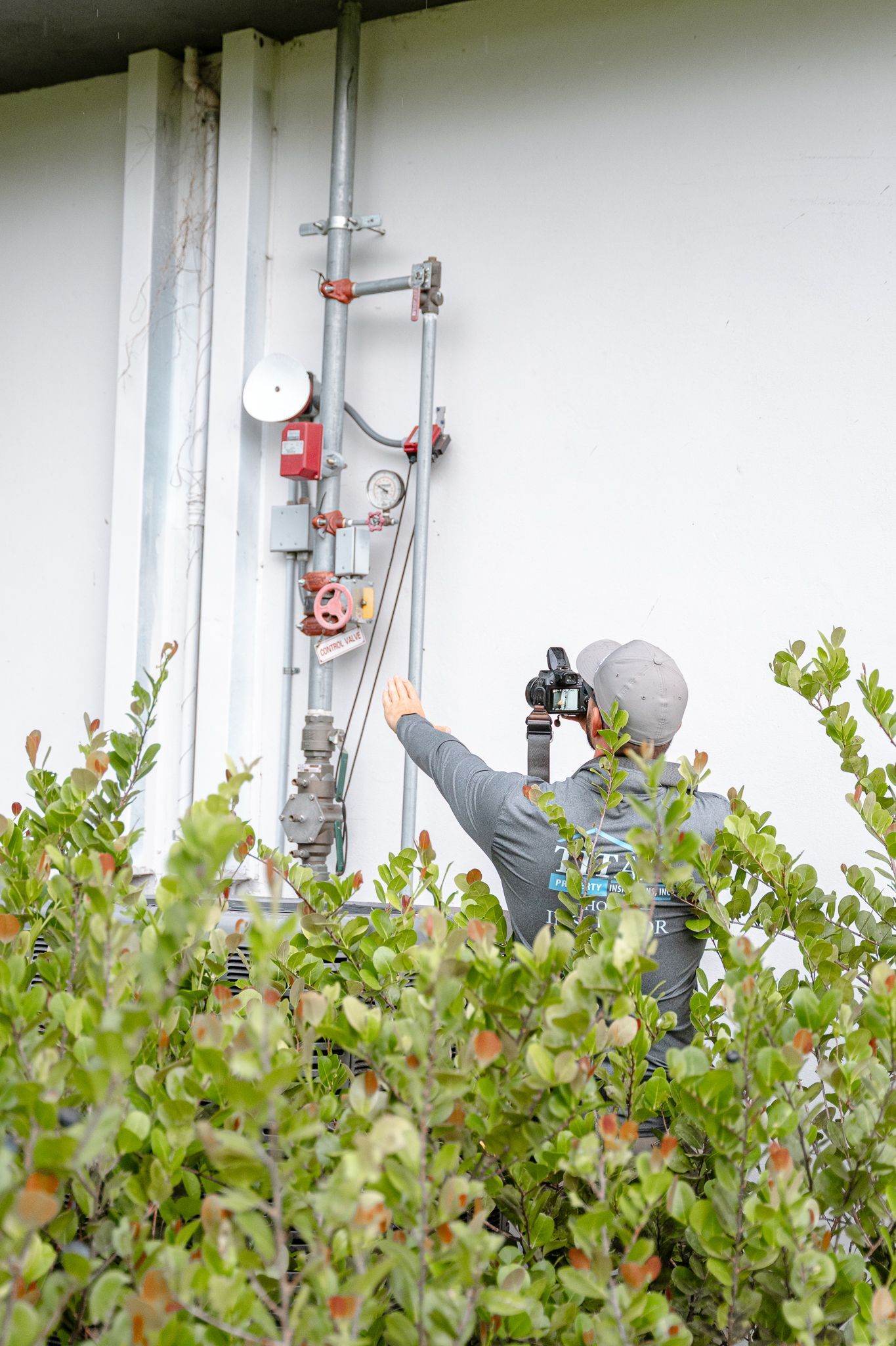 Persona señala tuberías y equipos en el exterior de un edificio blanco, oculto por arbustos verdes.