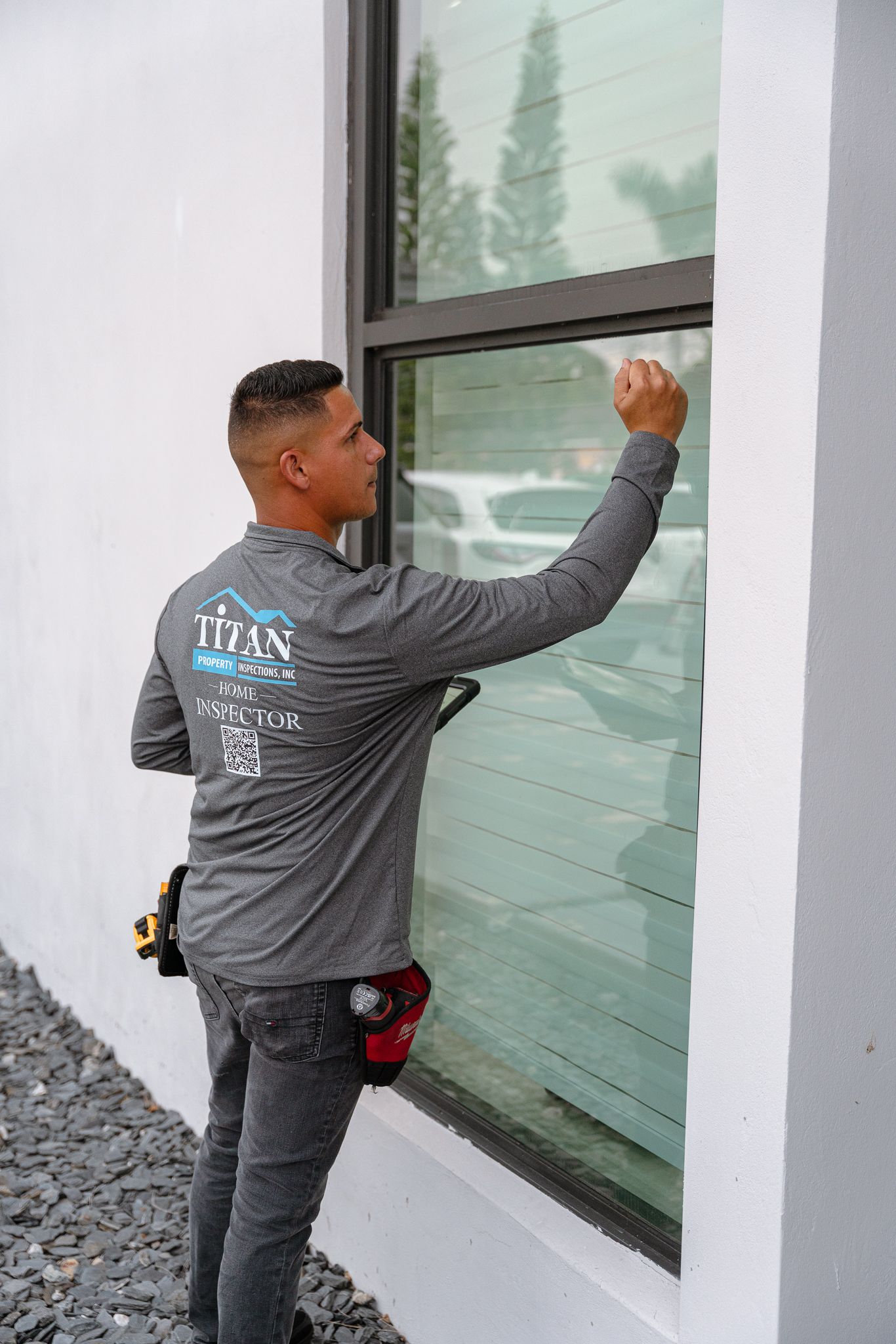 Hombre aplicando tinte a una ventana de un edificio blanco. Viste uniforme de trabajo gris con un logotipo y lleva un cinturón de herramientas.