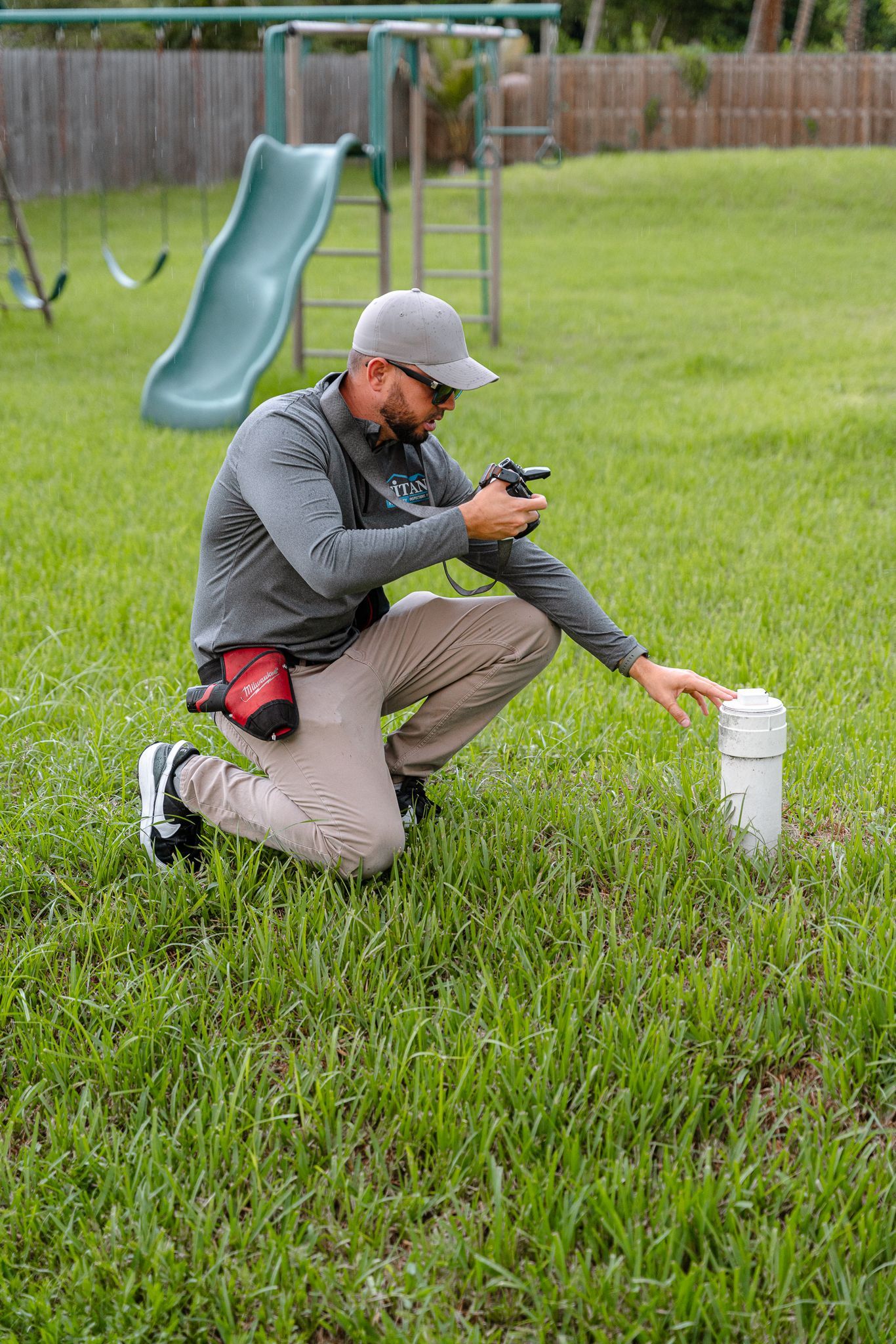 Un hombre se arrodilla en el patio, tocando una tubería y mirando un aparato. Al fondo se ve un parque infantil.