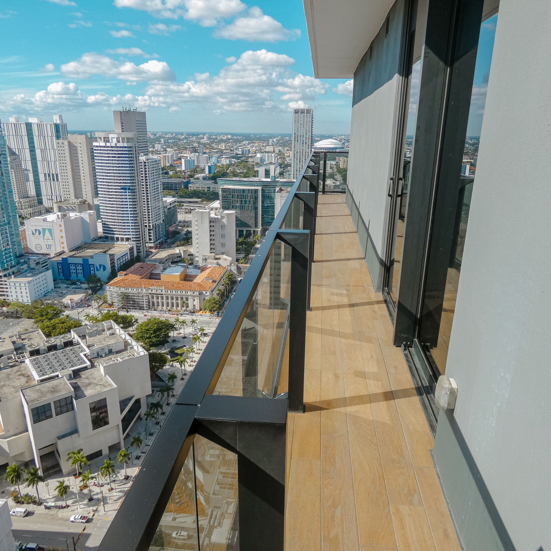 Vista panorámica de la ciudad desde un balcón con barandillas de cristal y suelo de madera. Día soleado y luminoso.