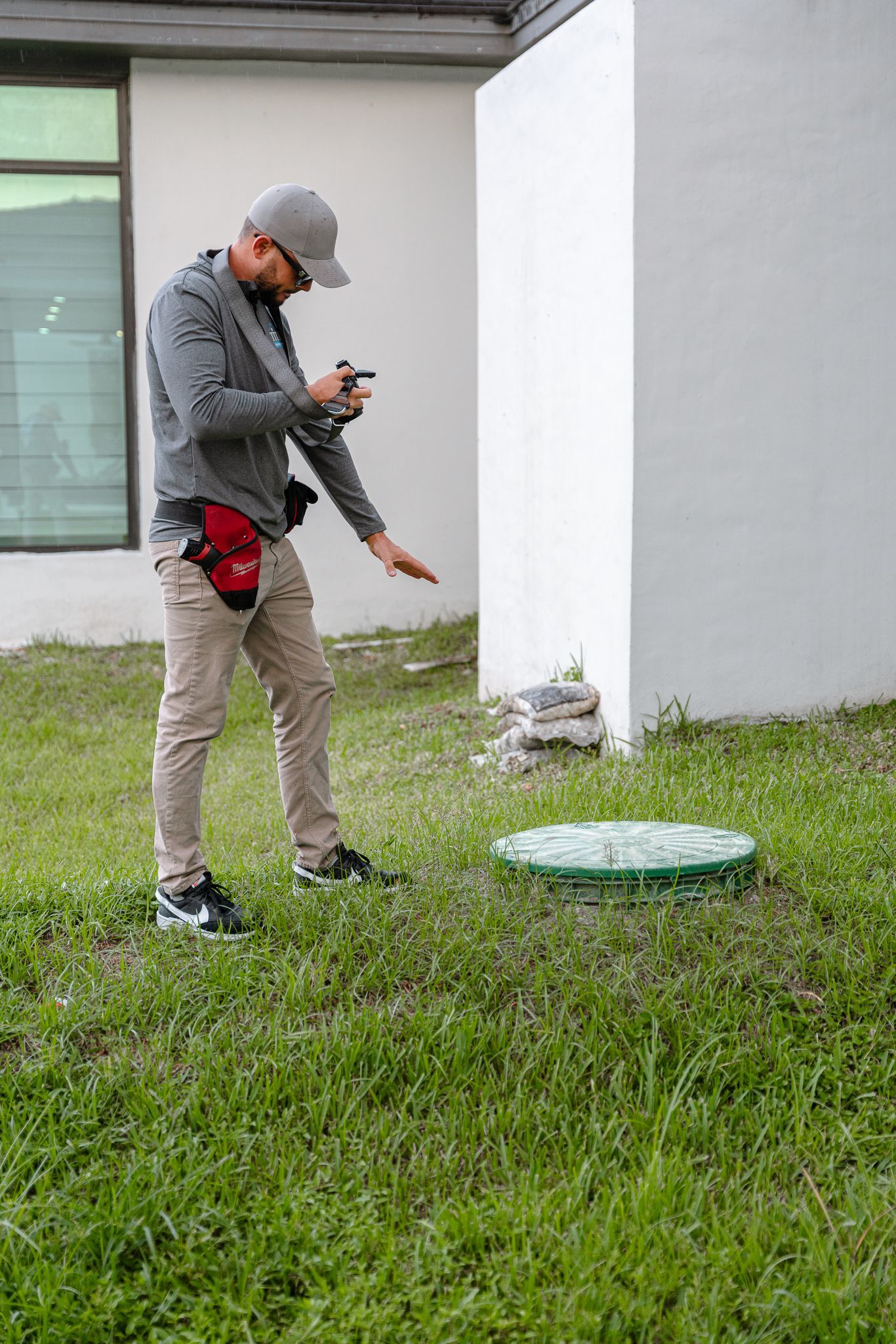 Hombre inspeccionando una tapa de servicio verde en un césped cerca de un edificio blanco.