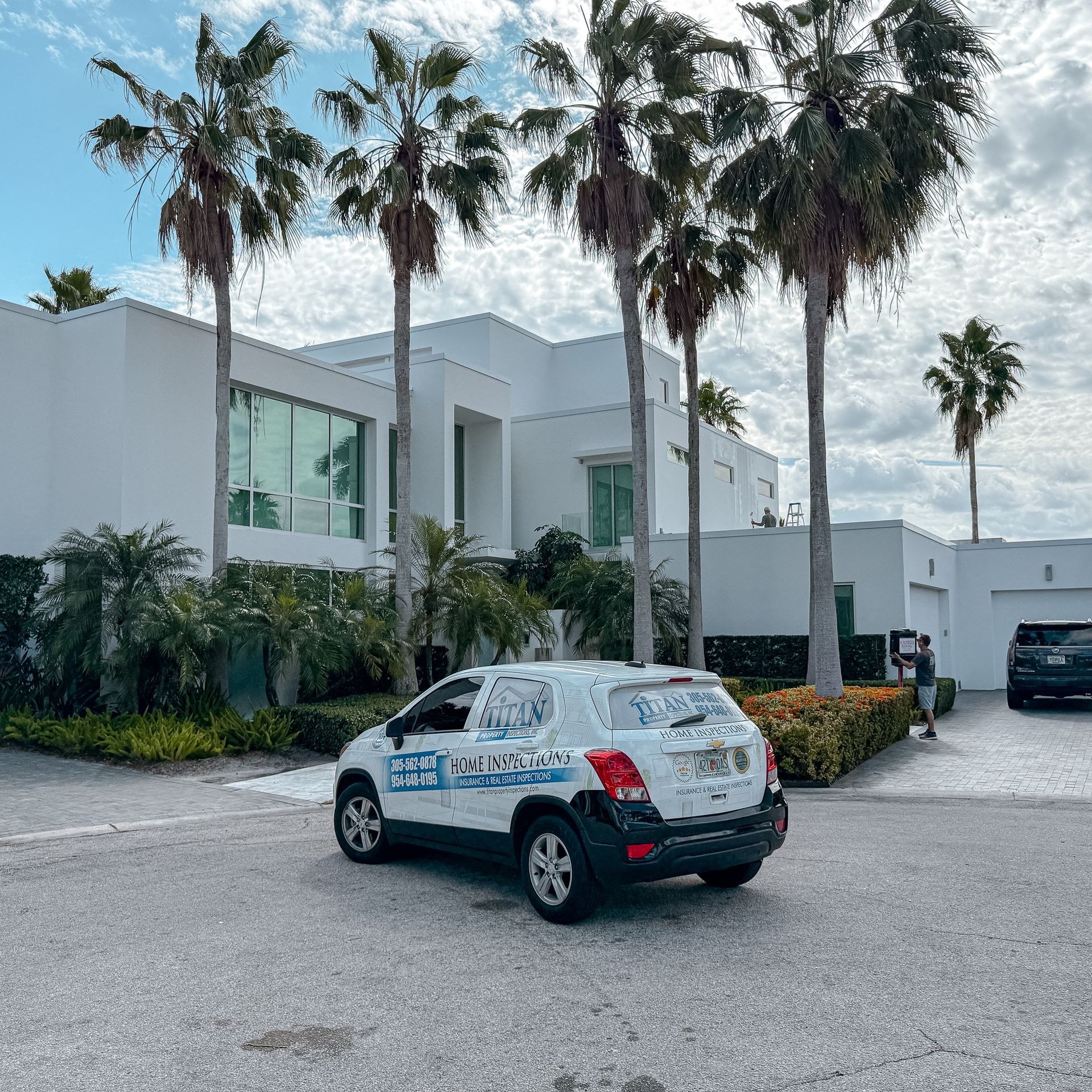 SUV blanco estacionado frente a una casa blanca moderna con palmeras; cielo nublado.