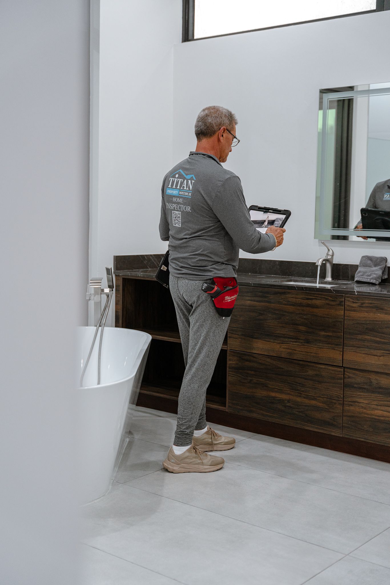 Hombre vestido de gris inspeccionando un baño moderno con bañera y tocador.