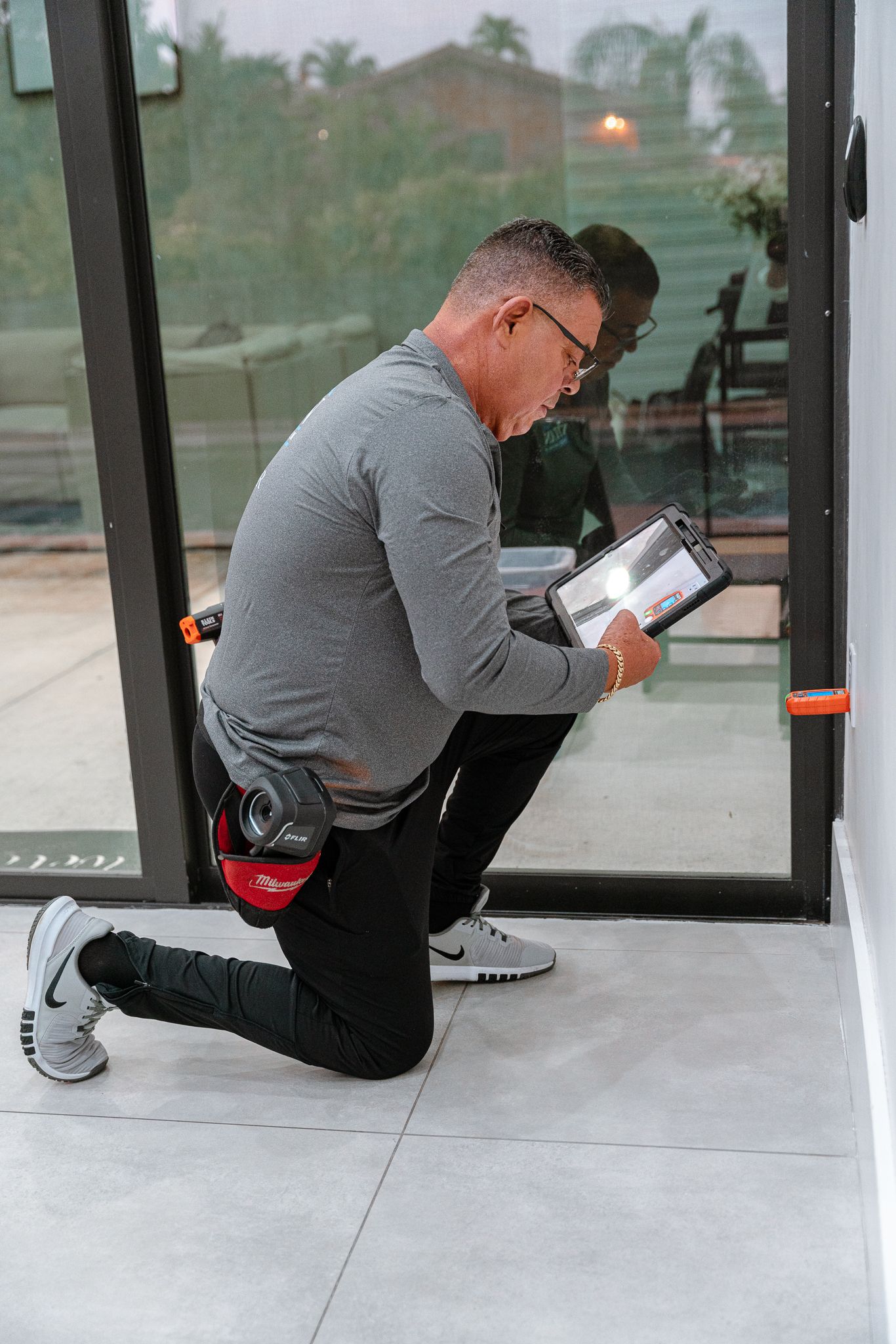 Una persona se arrodilla, inspeccionando una puerta. Lleva gafas, camisa gris, pantalón negro y zapatillas deportivas.