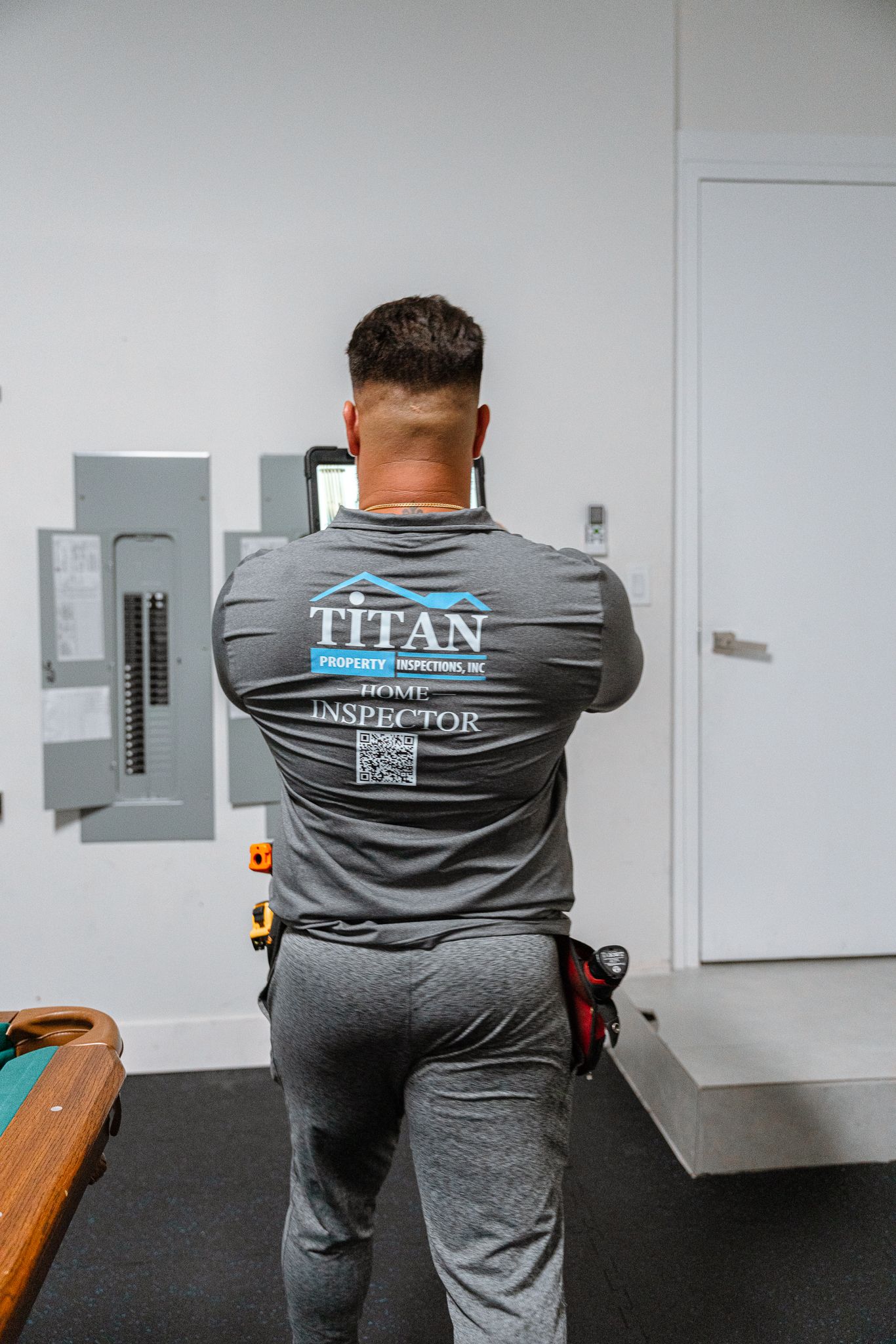 Un hombre con una camisa gris Titan Electric se encuentra frente a un panel eléctrico, con los brazos cruzados.