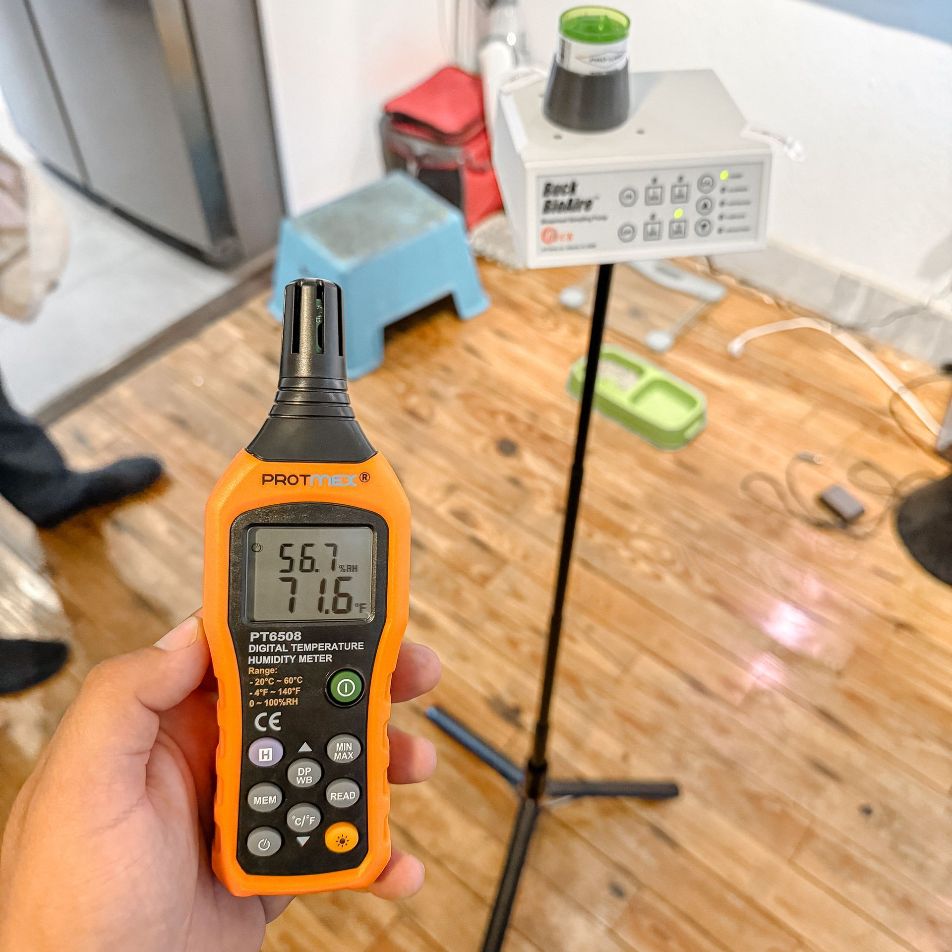 Una mano sostiene un medidor de calidad del aire naranja, con lecturas del 56,7 % y del 71,6 %. Hay otro medidor de calidad del aire en un soporte en la habitación.