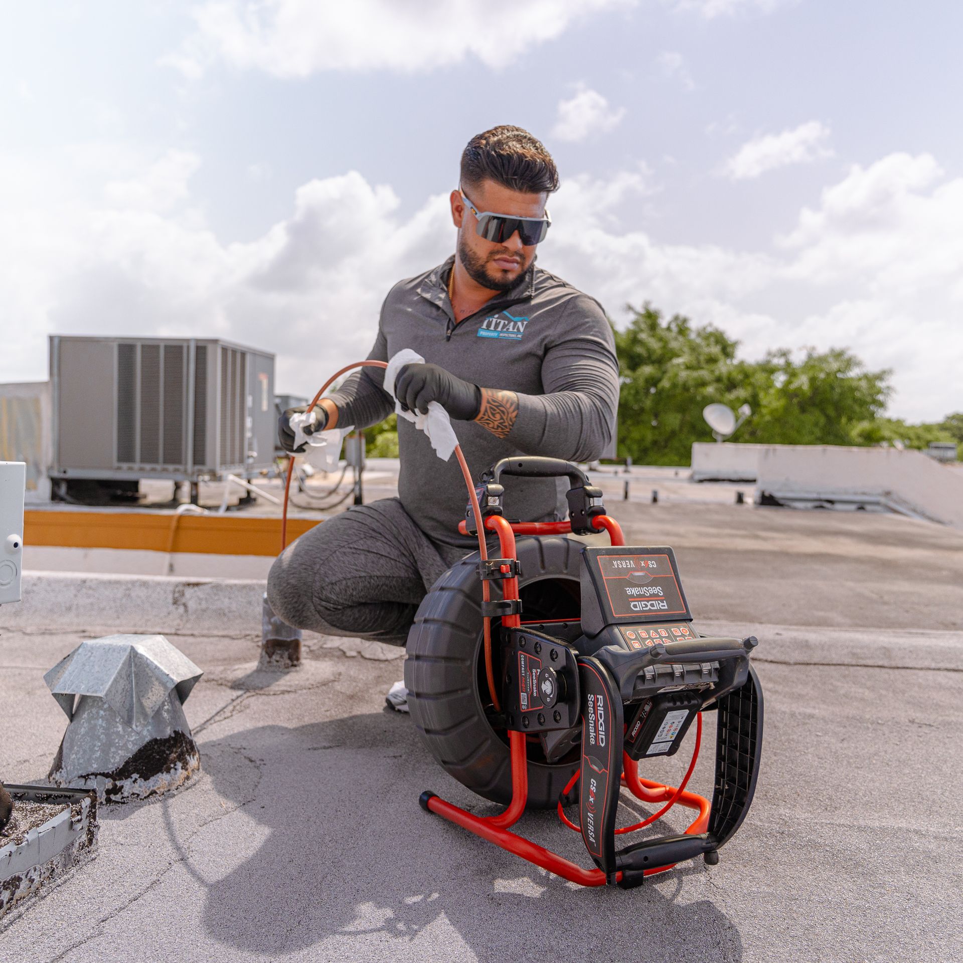 Hombre usando una cámara de alcantarillado en una azotea; mono gris, gafas de sol, guantes negros, sol brillante.