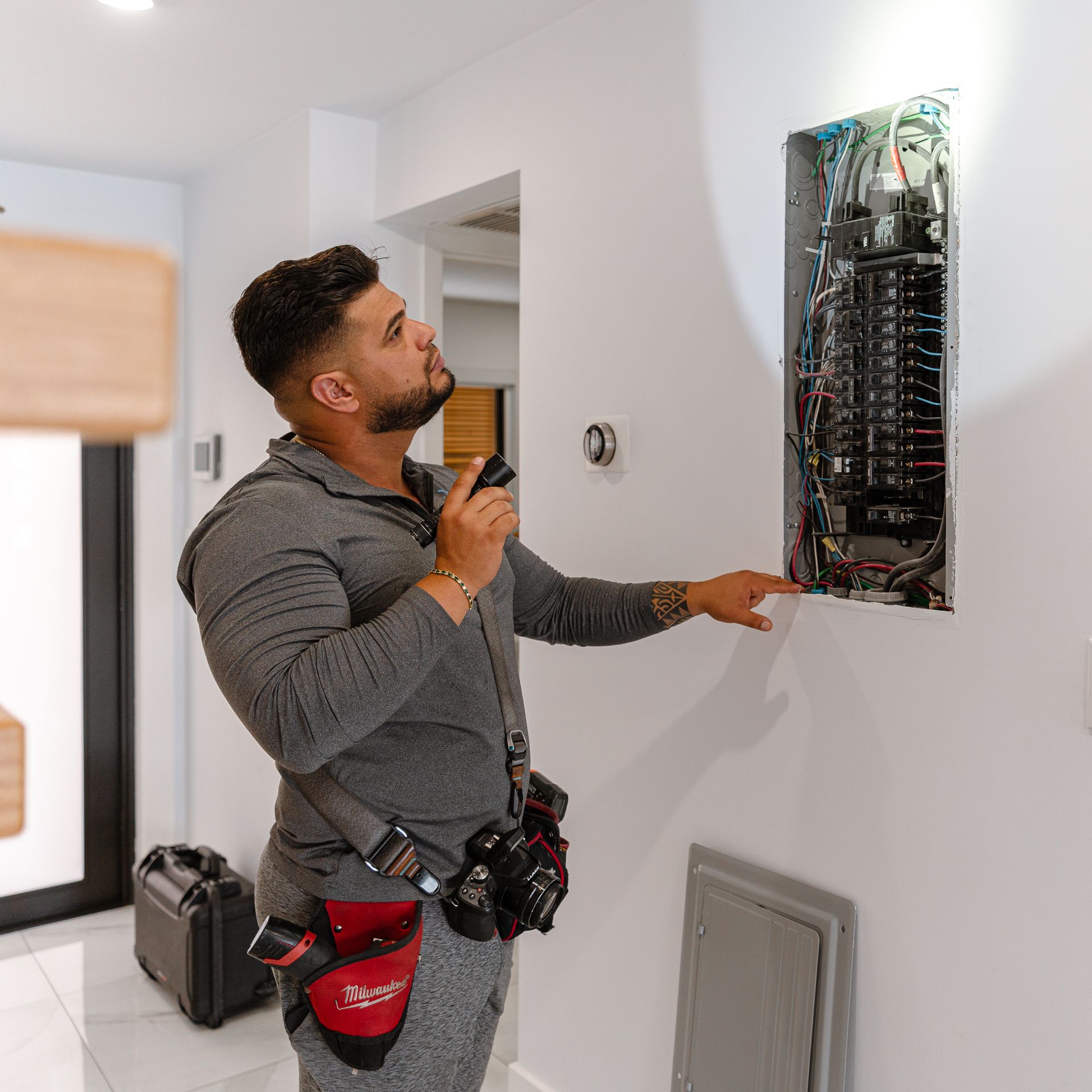 Un electricista examina un panel eléctrico con una linterna en la mano. Está en una habitación de paredes blancas y lleva un cinturón de herramientas.