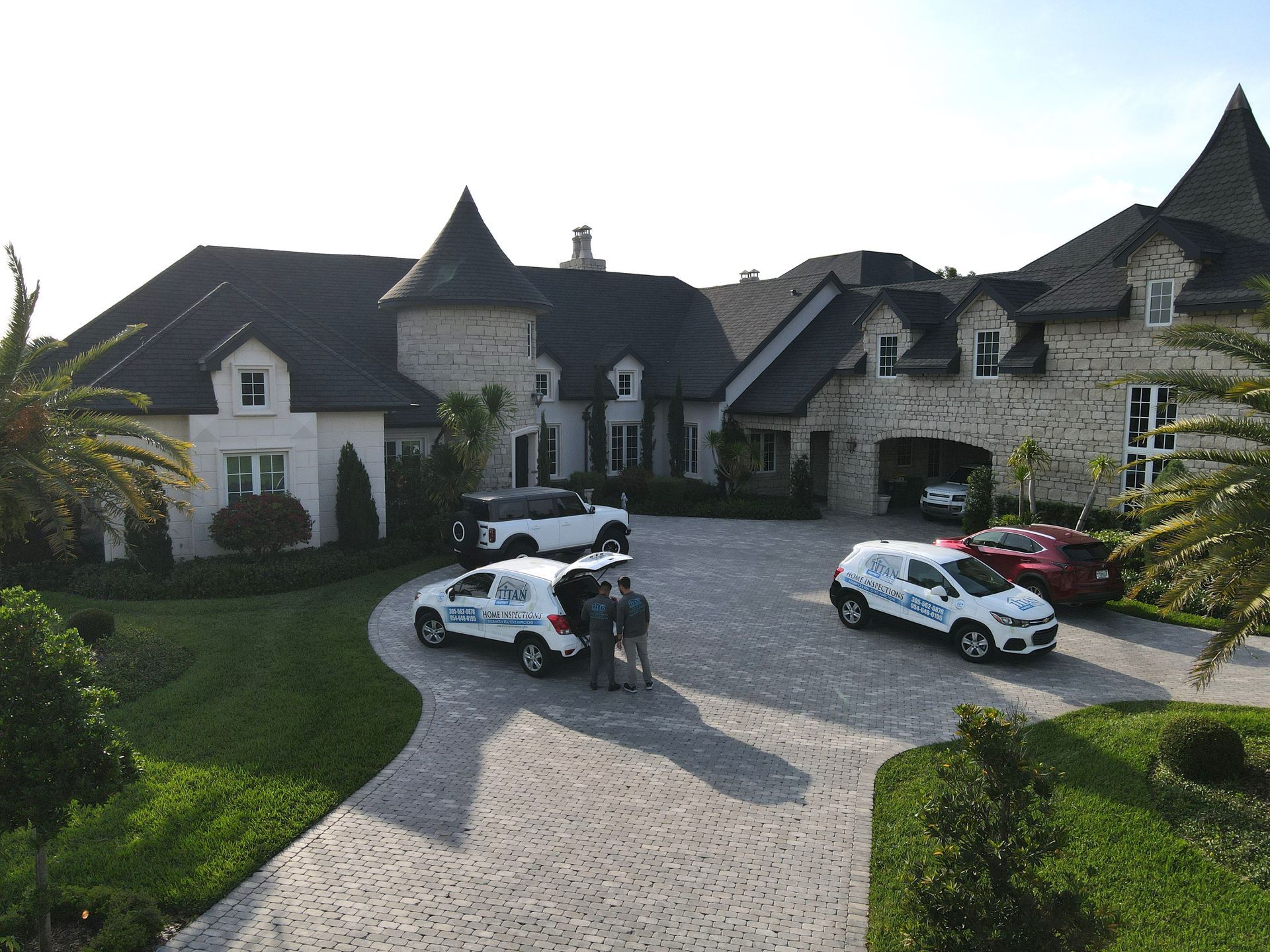 Coches aparcados delante de una gran casa de piedra con torretas y jardines bien cuidados; dos personas están cerca de un vehículo de servicio.