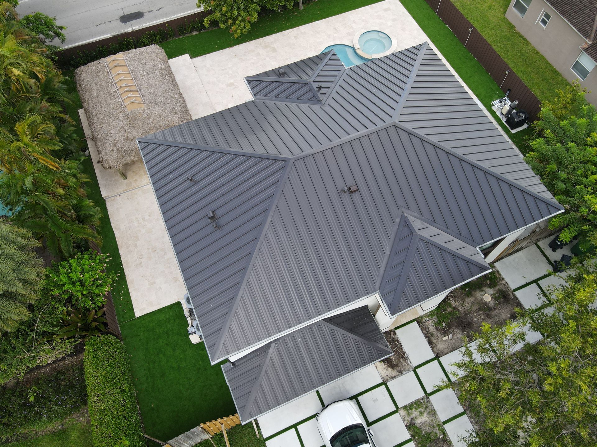 Vista aérea de una casa con techo de metal gris oscuro, una piscina y una cabaña tiki en un patio verde.
