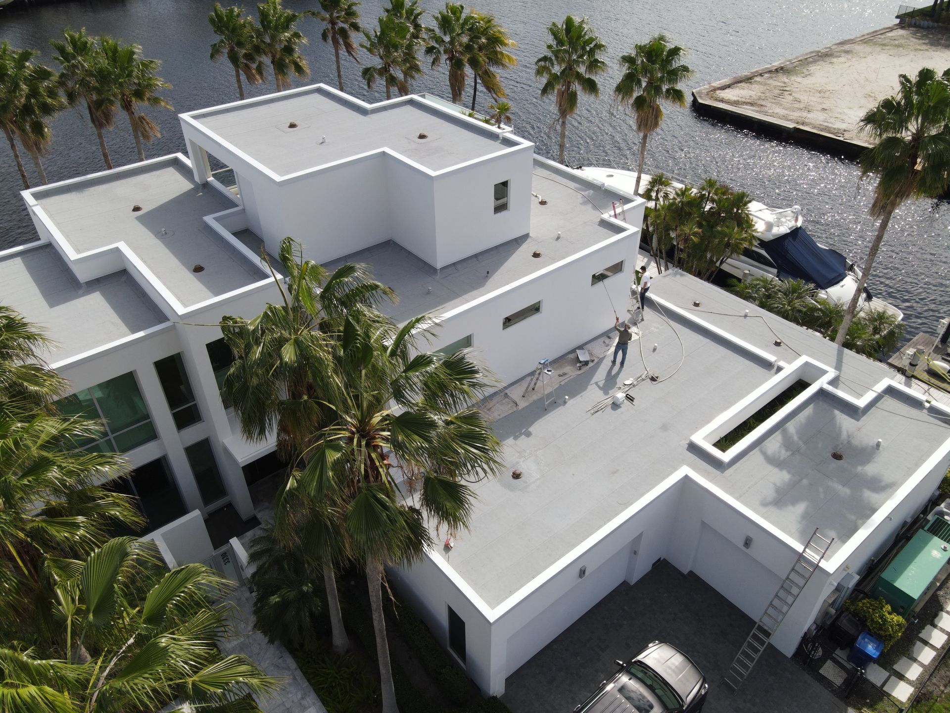 Moderna casa blanca con techo plano junto al agua, palmeras y un auto en la entrada.