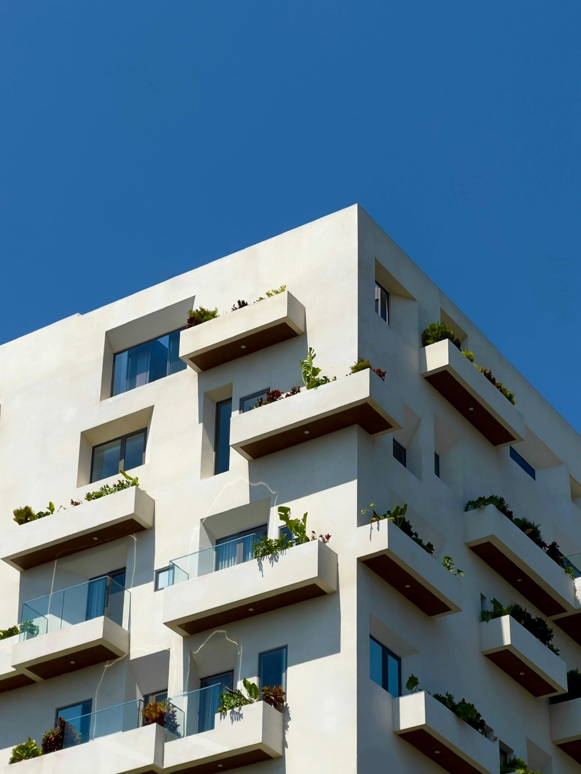 Edificio moderno blanco con balcones angulares, exuberante vegetación y cielo azul.