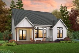 Small white house with gray roof, lit windows, front porch, and landscaped yard at sunset