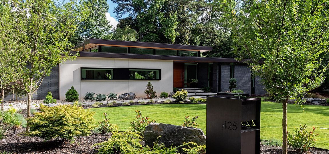 Modern single-story house with a large window, surrounded by trees and a landscaped garden