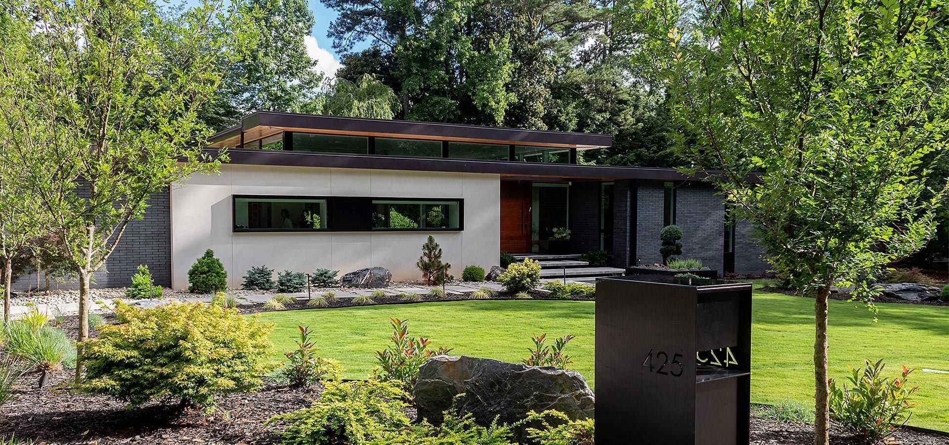 Modern single-story house with a large window, surrounded by trees and a landscaped garden