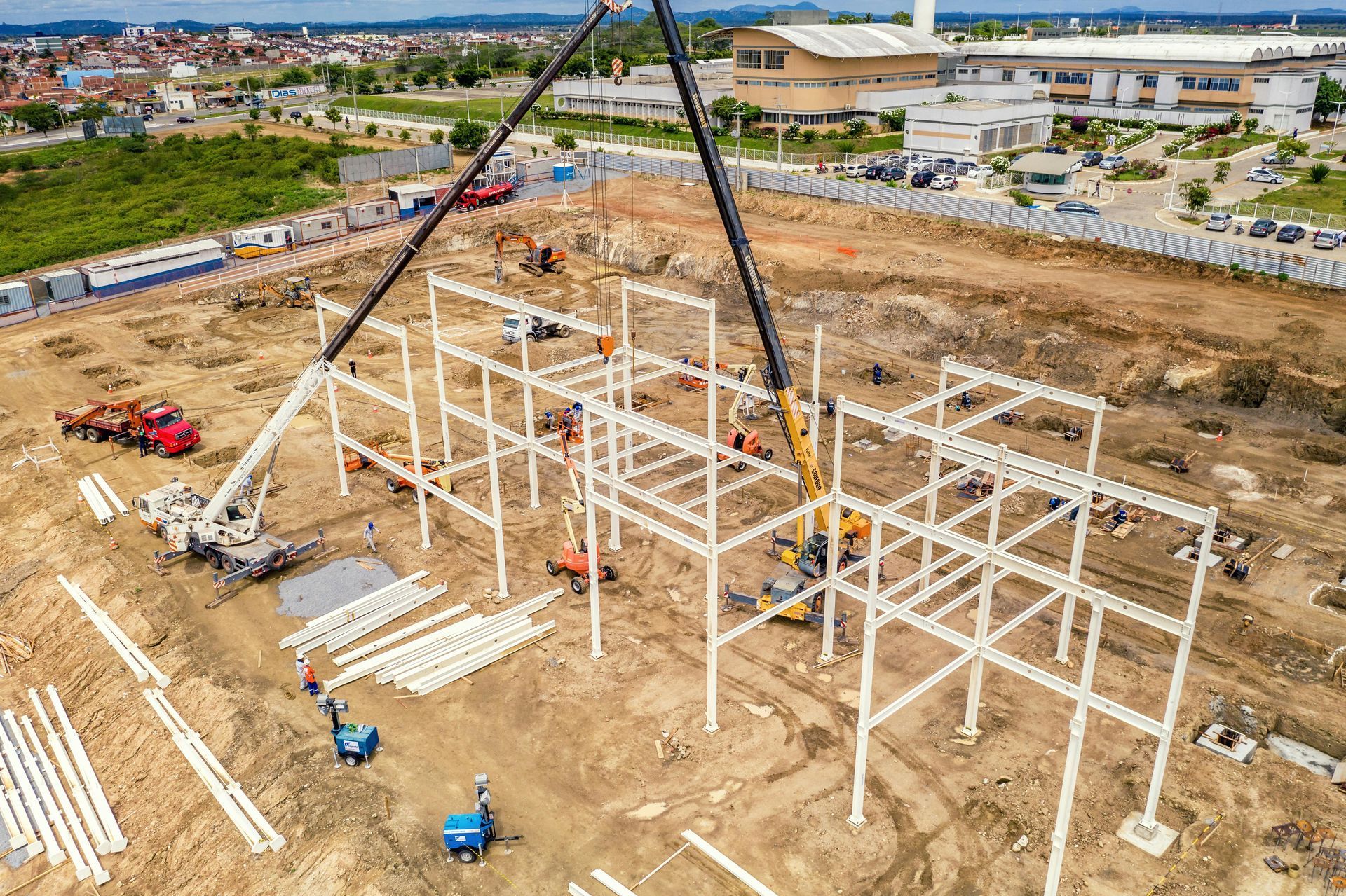 Construction site: crane lifting steel framework, workers on the ground, dirt, and buildings in the background.