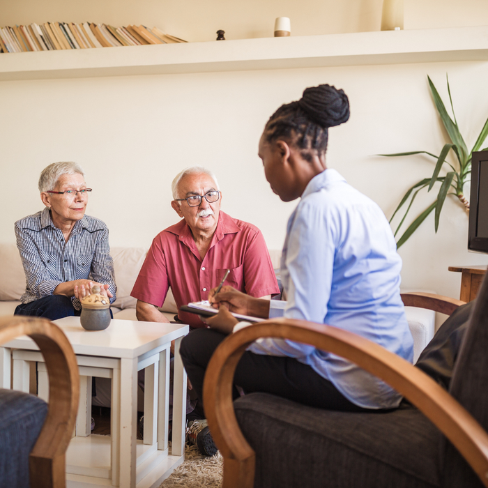 A healthcare professional sitting in a chair, taking notes while talking to two individuals seated on a sofa.