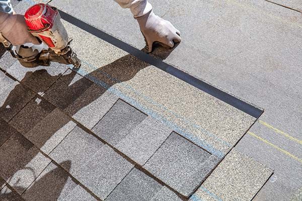 Roofer installing asphalt shingles with a nail gun on a rooftop in daylight.