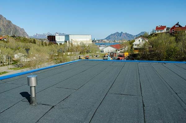A flat, dark gray roof with a metal vent, overlooking a town and mountains under a blue sky.