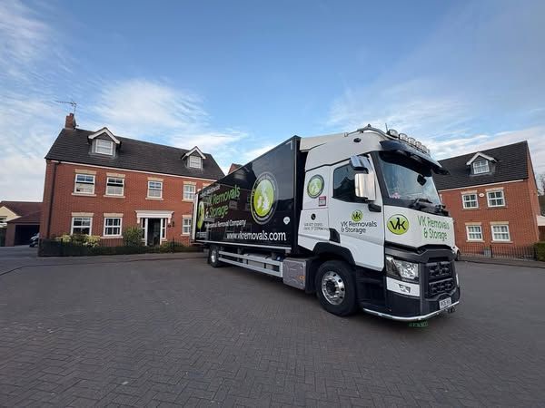Three people pose with a VK Removals truck in front of houses on a sunny day.