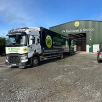 A white and blue VK Removals truck parked outside a green warehouse.