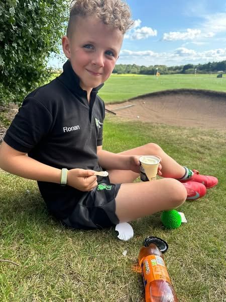Boy in black shirt and shorts, eating ice cream on golf course, sunny day.