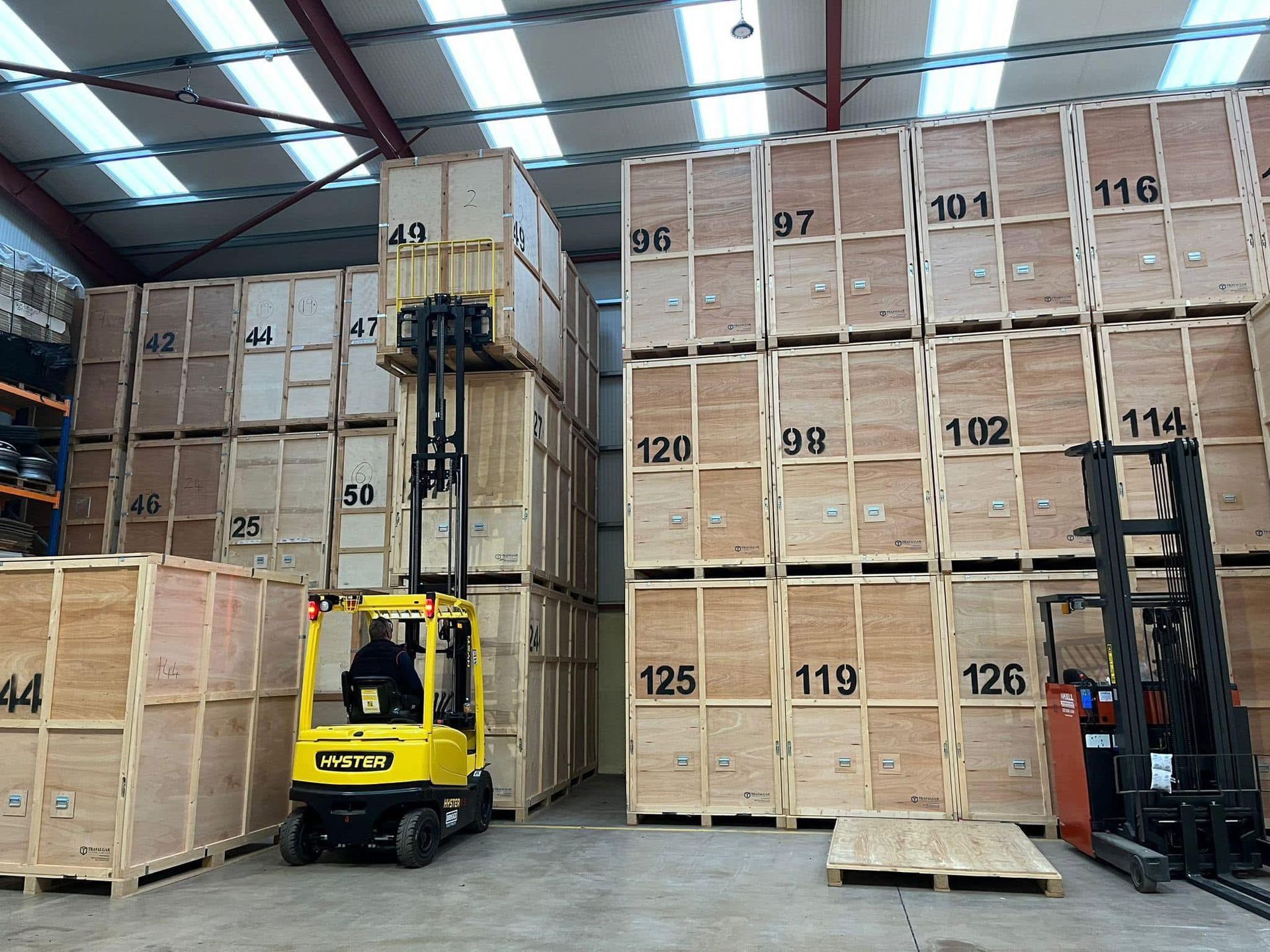 A yellow forklift stacks wooden crates in a warehouse. Crates are labeled with numbers.