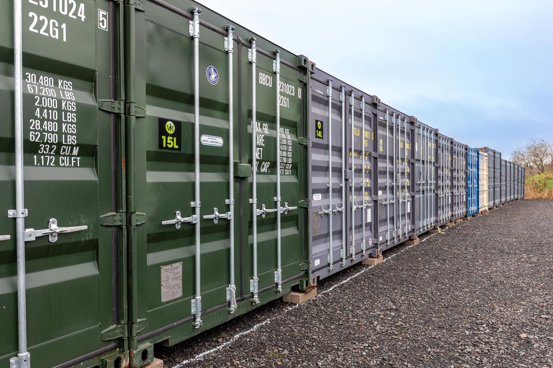 Green and gray shipping containers lined up in a row on gravel under a cloudy sky.