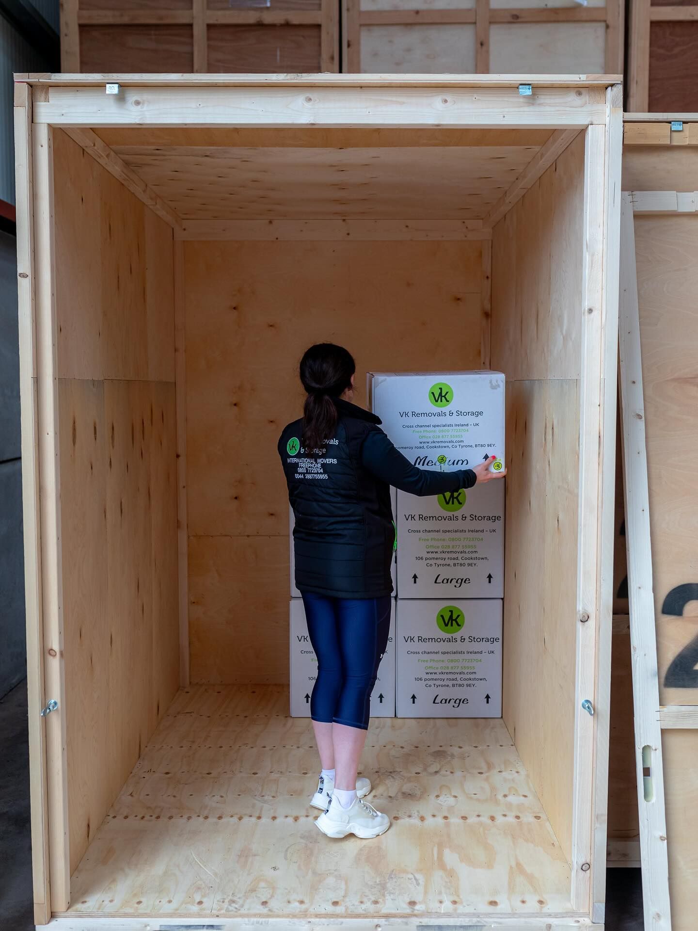 Woman loading boxes into a wooden storage unit, wearing black jacket and blue leggings.