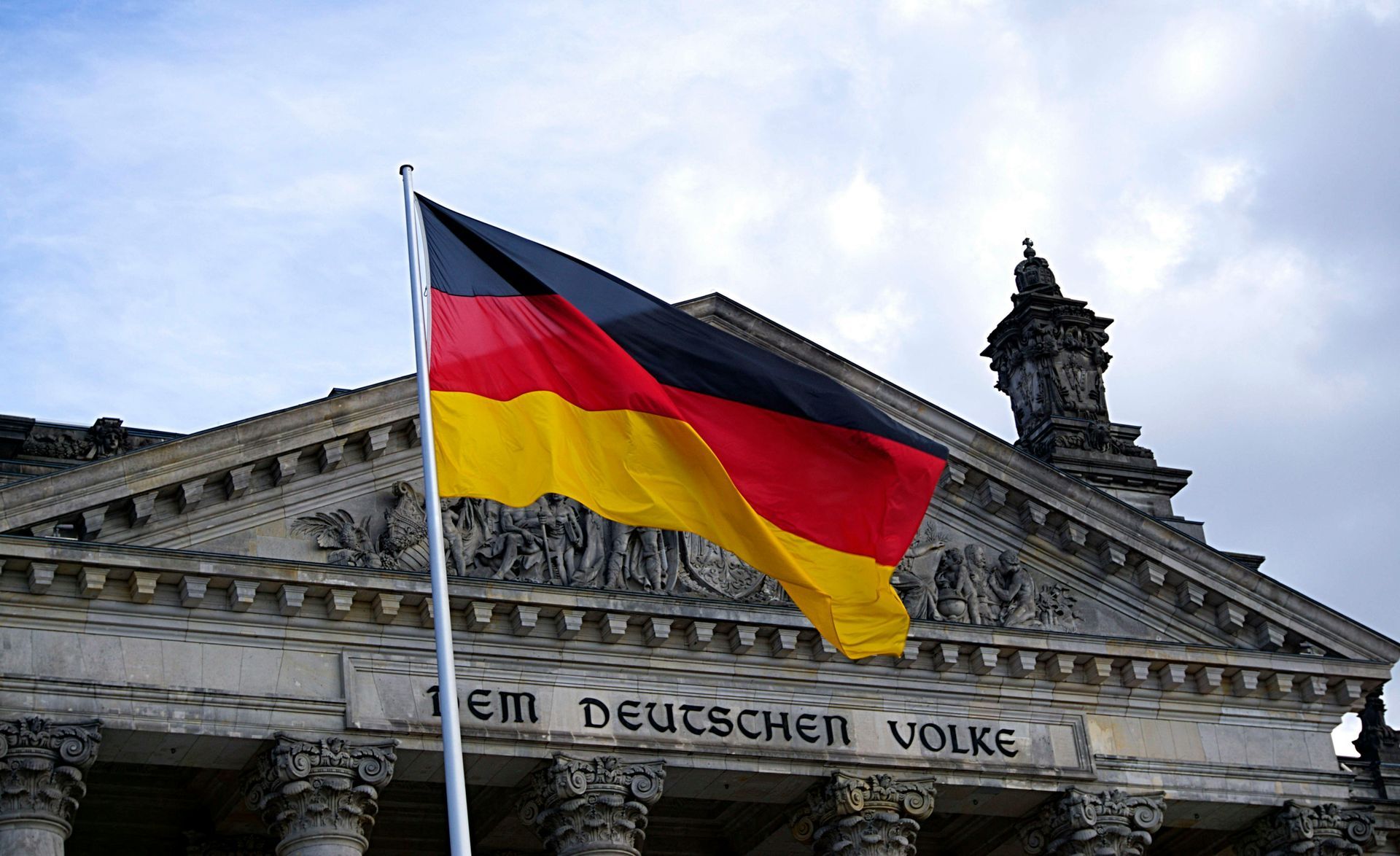 German flag waving in front of the Reichstag building, displaying black, red, and gold colors.