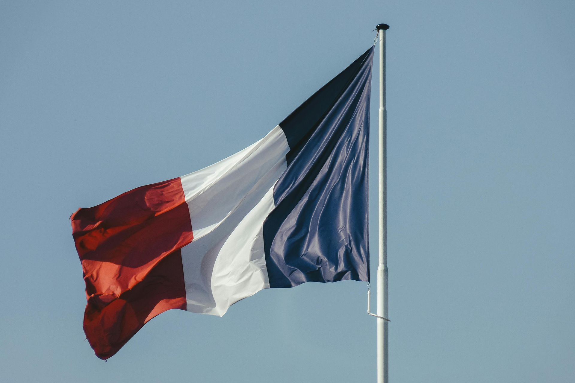 French flag waving on a flagpole against a blue sky, showing red, white, and blue vertical stripes.
