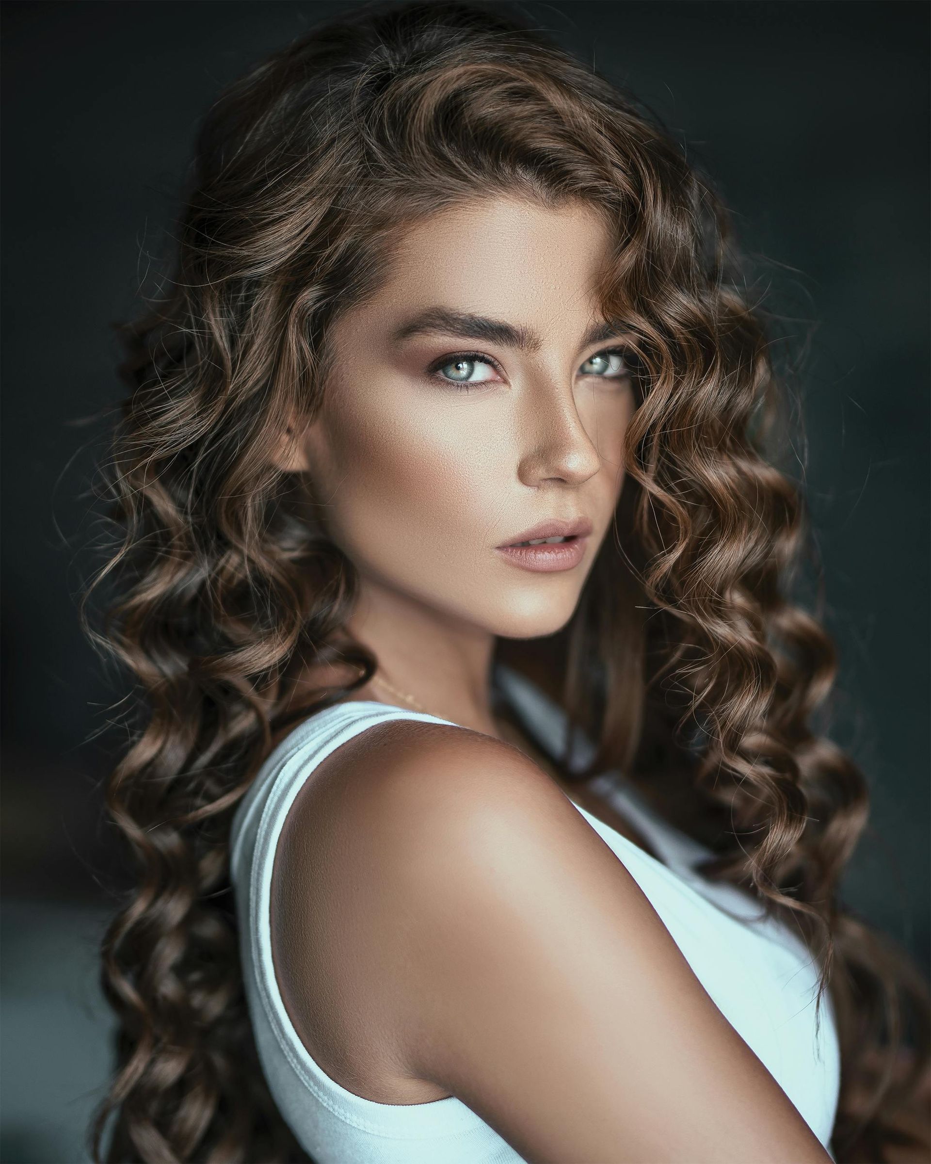 A woman with long curly hair and freckles is wearing a white tank top.