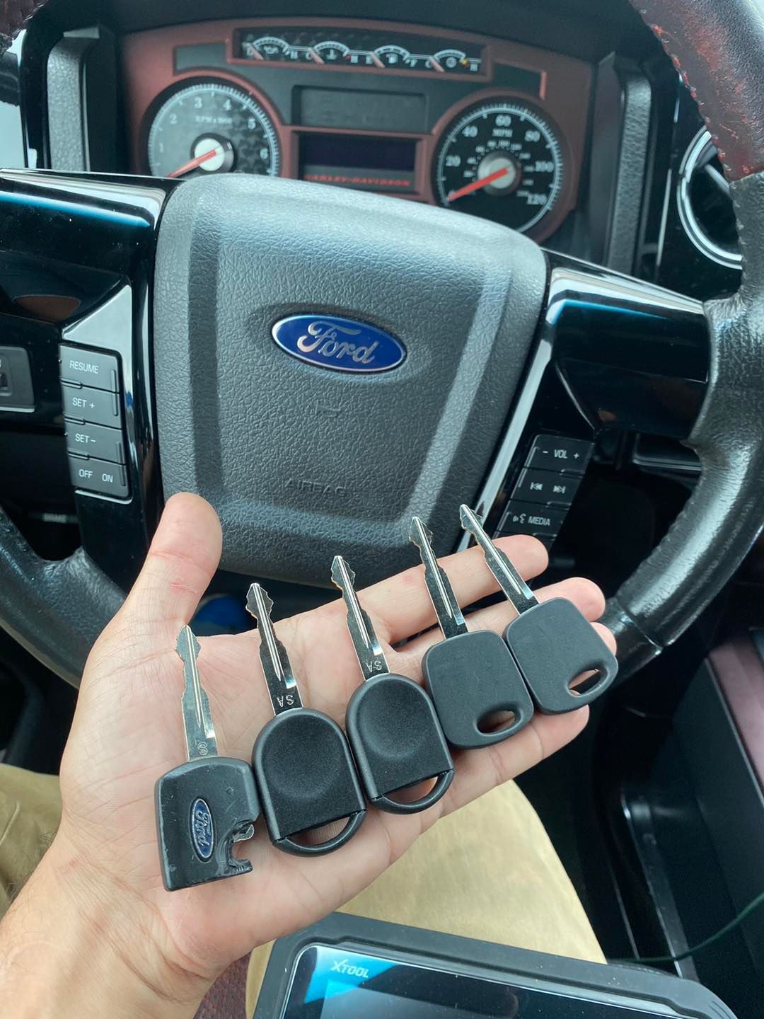 Hand holding five Ford keys in front of a Ford steering wheel, likely inside a vehicle.
