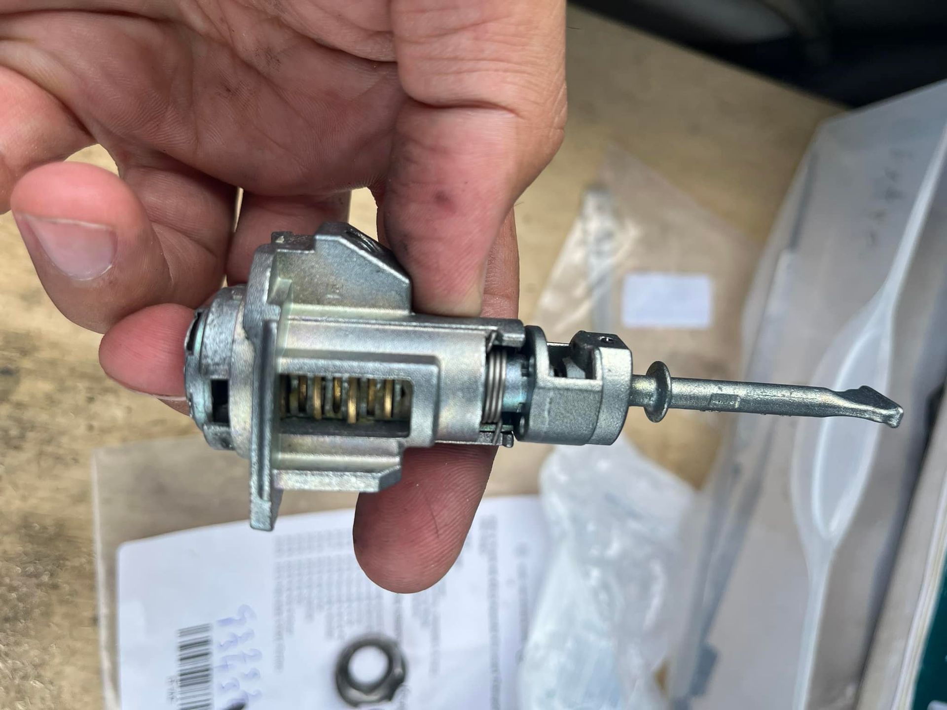 Hand holding a new car ignition lock cylinder. Silver metal, internal gears visible.