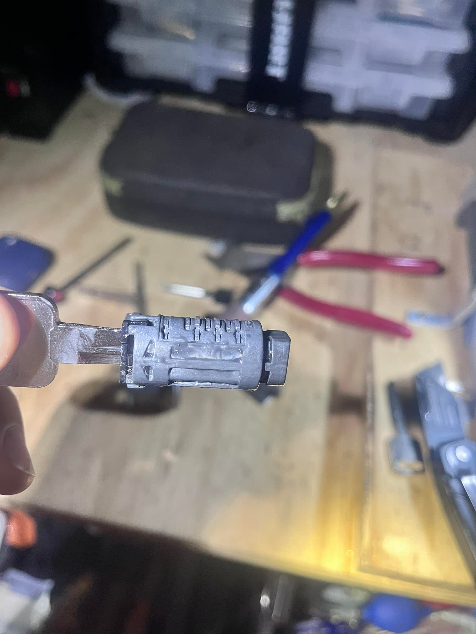 Close-up of a car ignition lock cylinder and key held by a hand; tools in the blurred background.