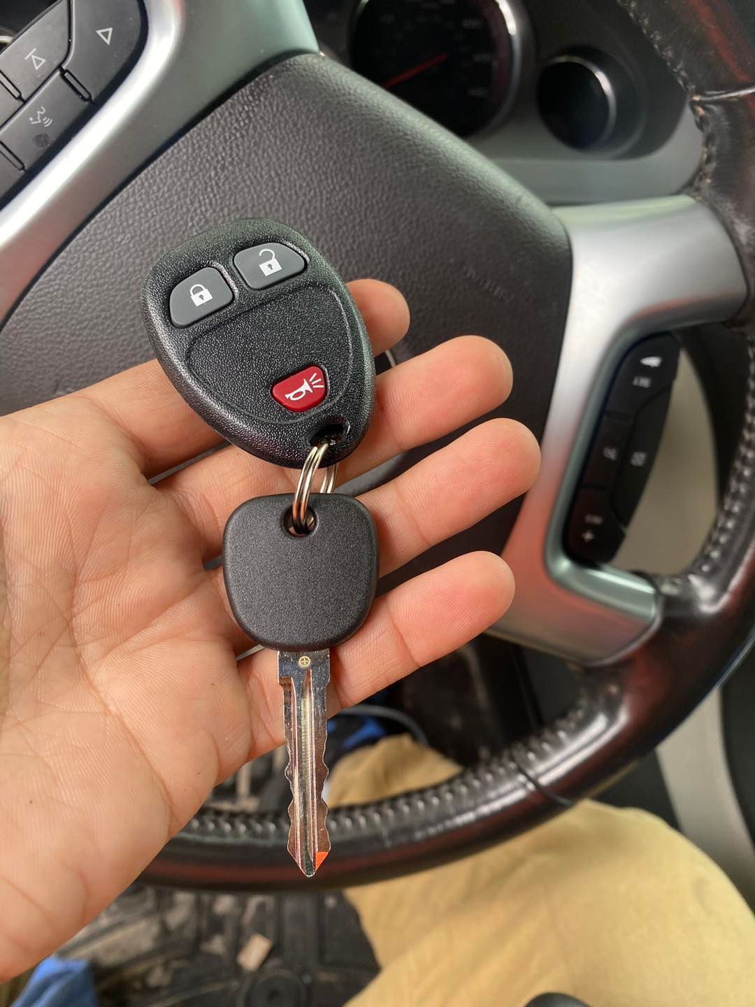 Hand holding car keys with remote, near a steering wheel. Black key, red button, silver accents.