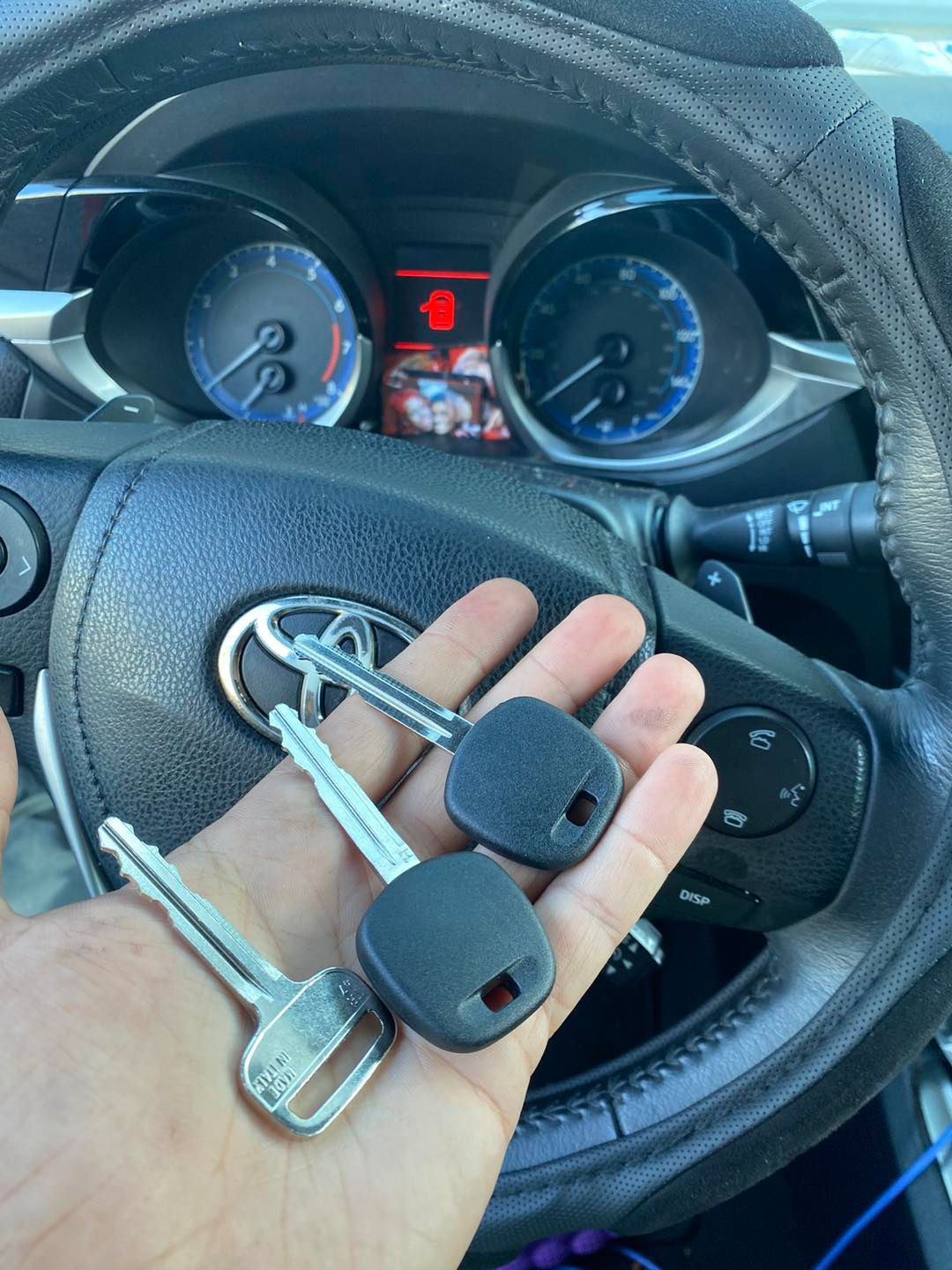 Hand holding three car keys in front of a Toyota steering wheel and dashboard.