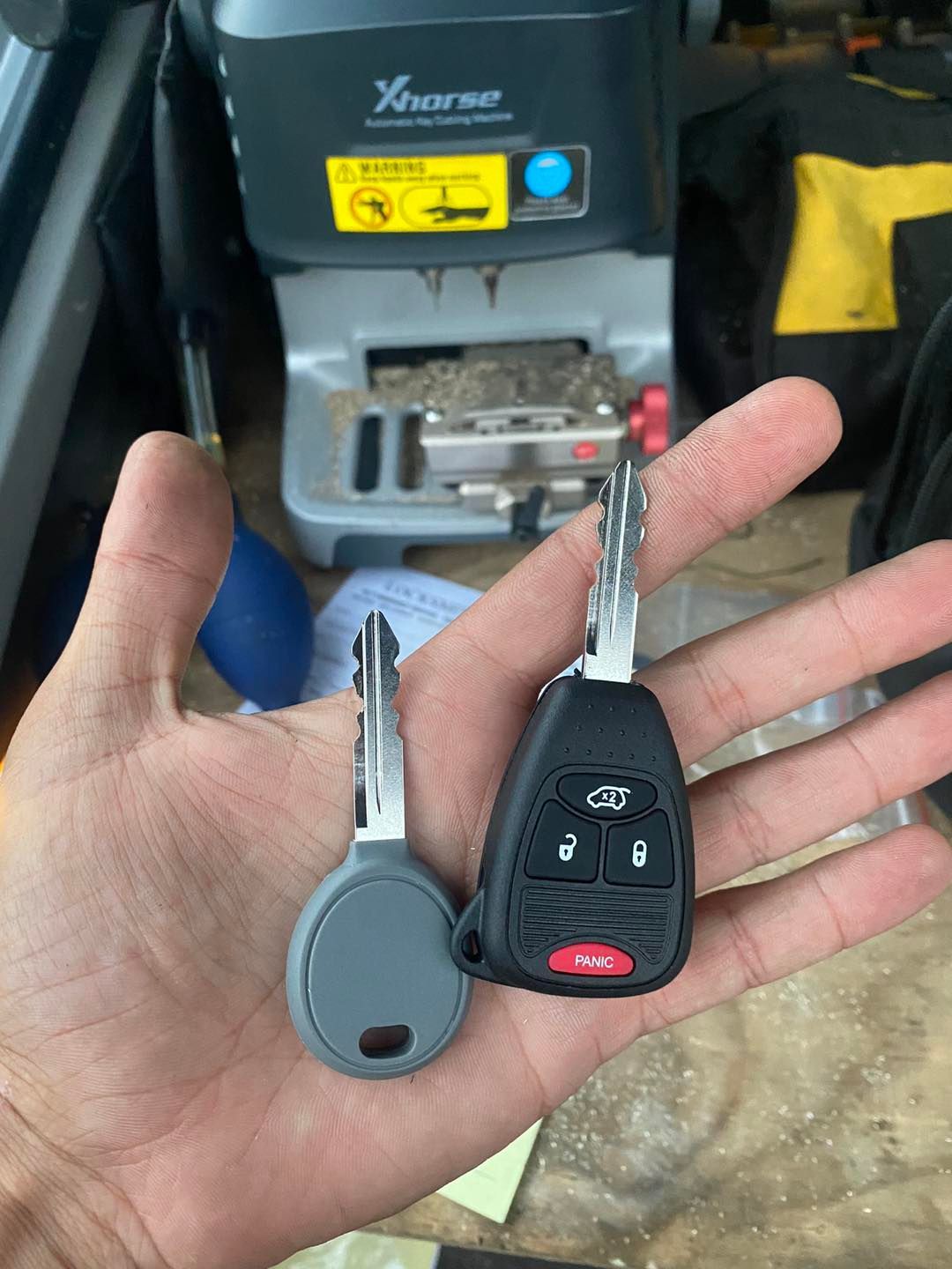 Hand holding two car keys; one gray, one black fob with red panic button, near a key cutting machine.