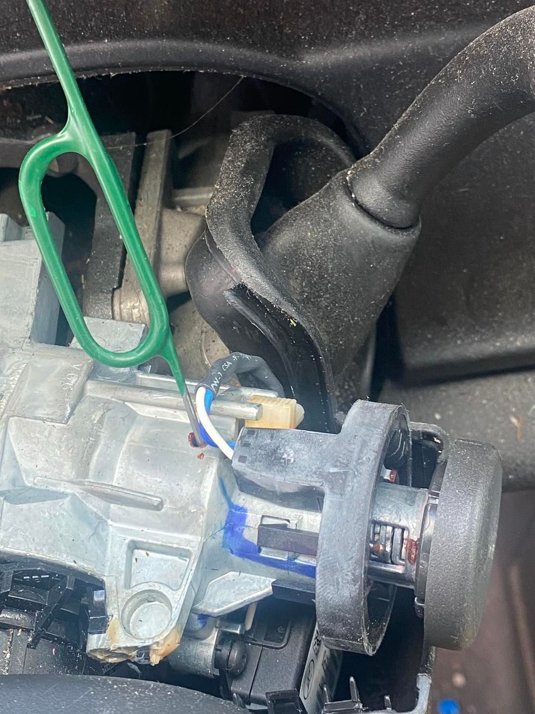 Close-up of a car ignition cylinder with wires attached, a green tool inserted, and a black steering column.