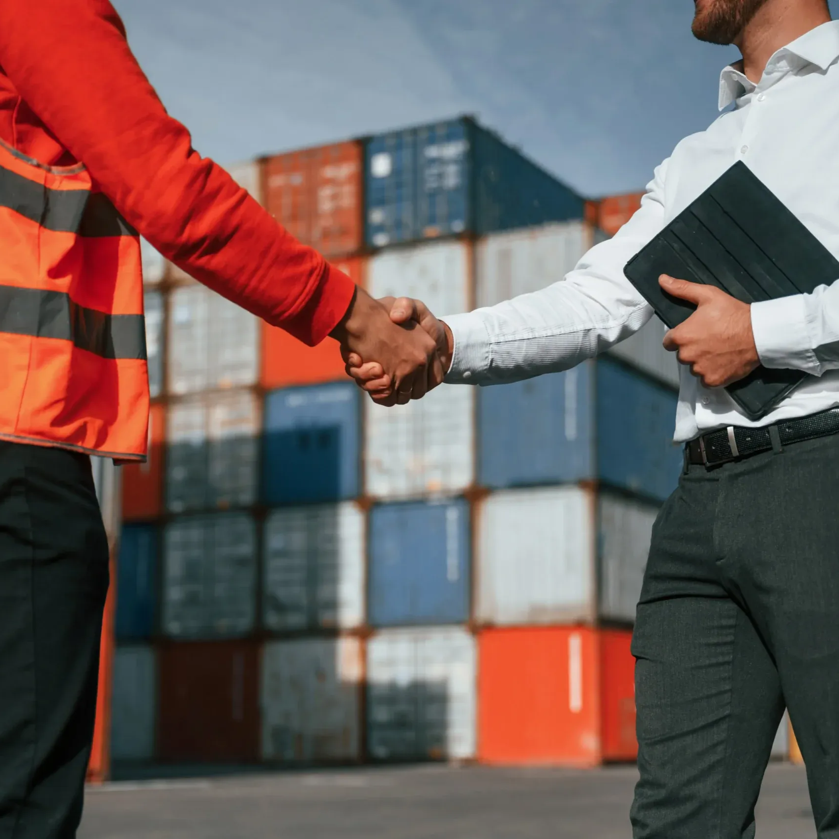 Two people shaking hands in front of cargo containers; one in a safety vest, the other holding a tablet.
