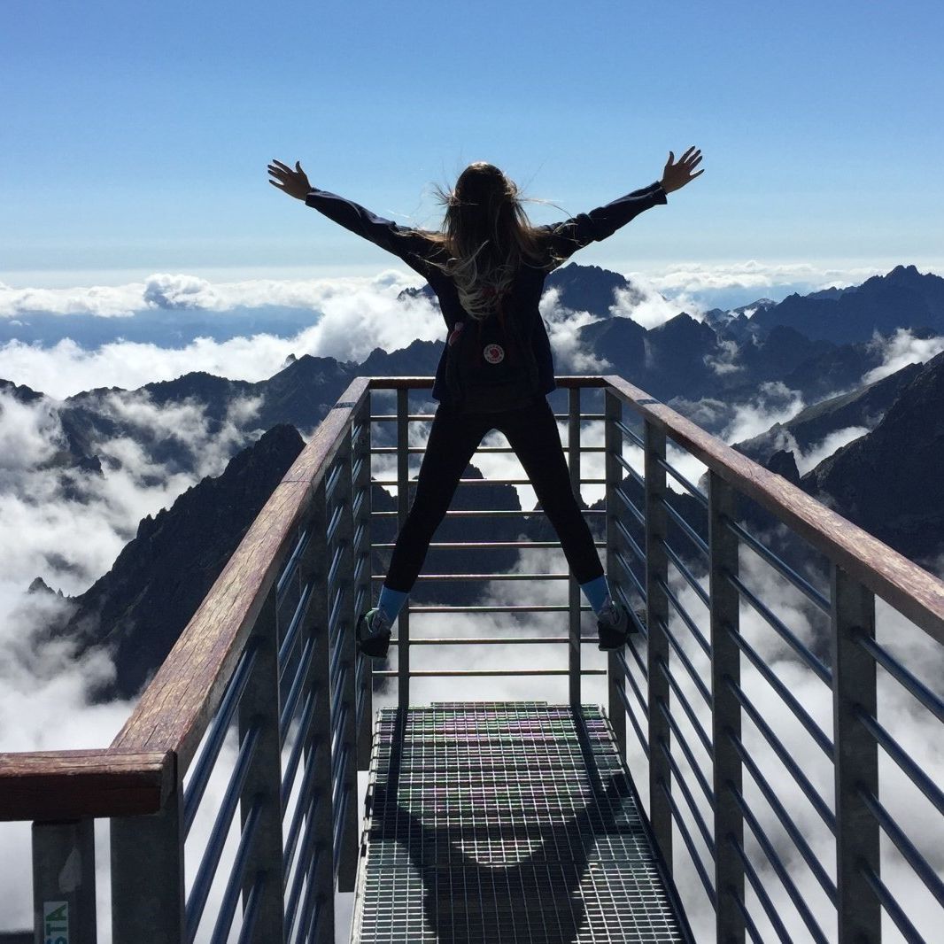 A woman is standing on a bridge overlooking mountains with her arms outstretched