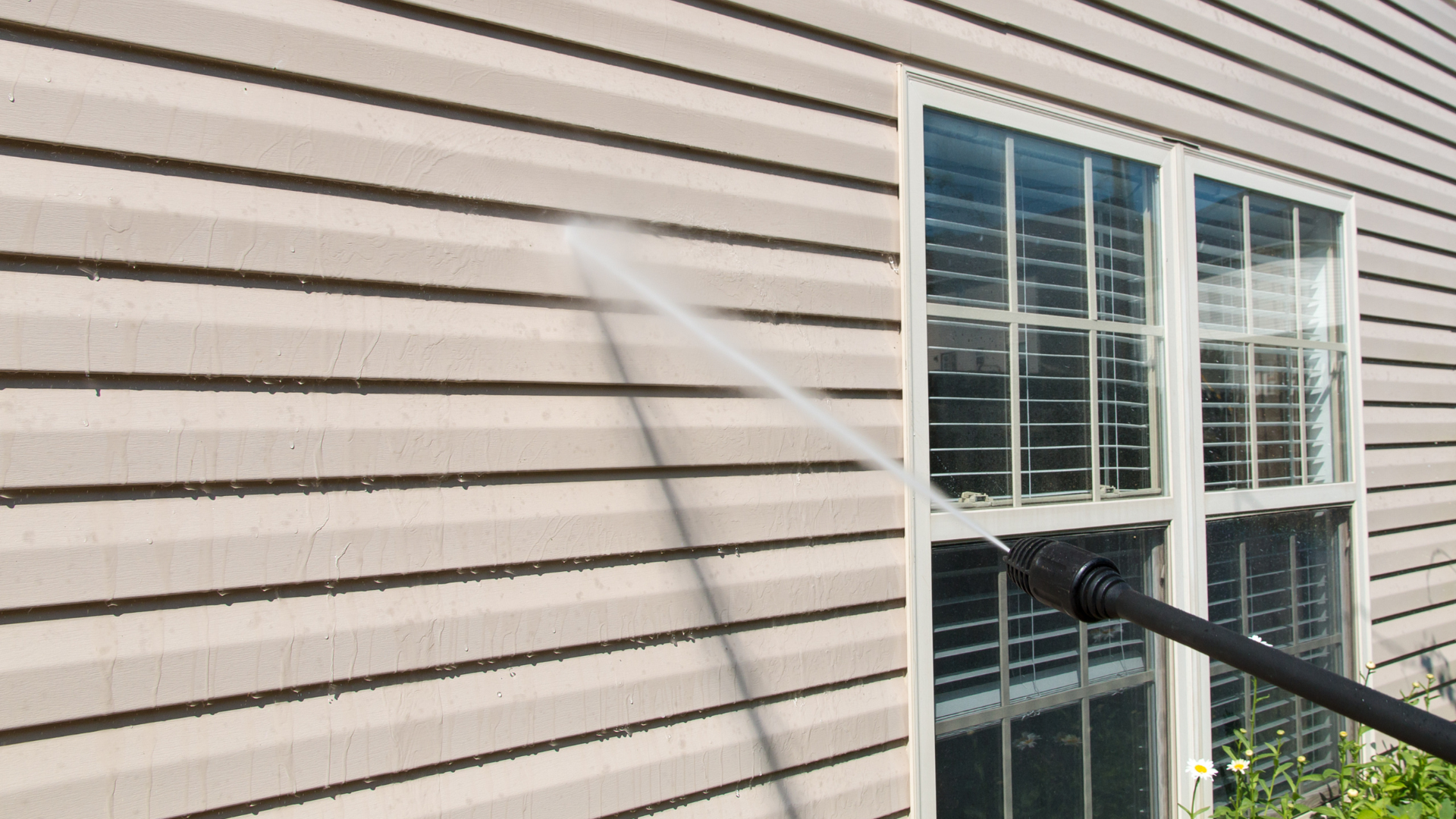 A power washer spray nozzle cleans a section of light-colored vinyl siding next to a white-framed window.