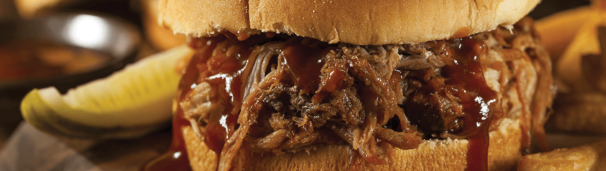 A close up of a pulled pork sandwich with a pickle on the side.