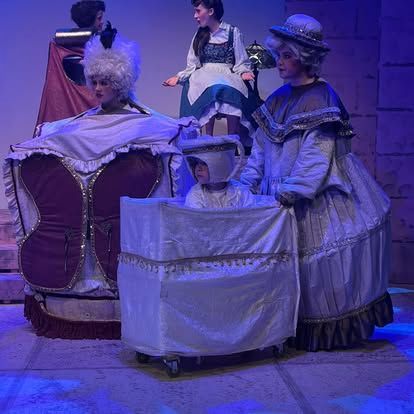 A group of women dressed in costumes are standing around a baby in a crib on a stage.
